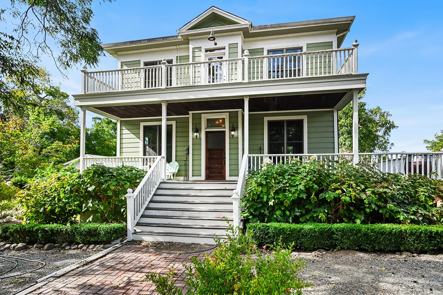 Front view of a two-story green house with white trim, a covered porch, a second-floor balcony, and front steps leading to the main entrance.