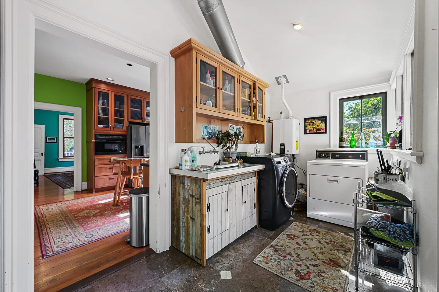 A laundry and utility room featuring a washer, dryer, sink cabinet, and upper wooden storage cupboards, leading into an adjacent kitchen.