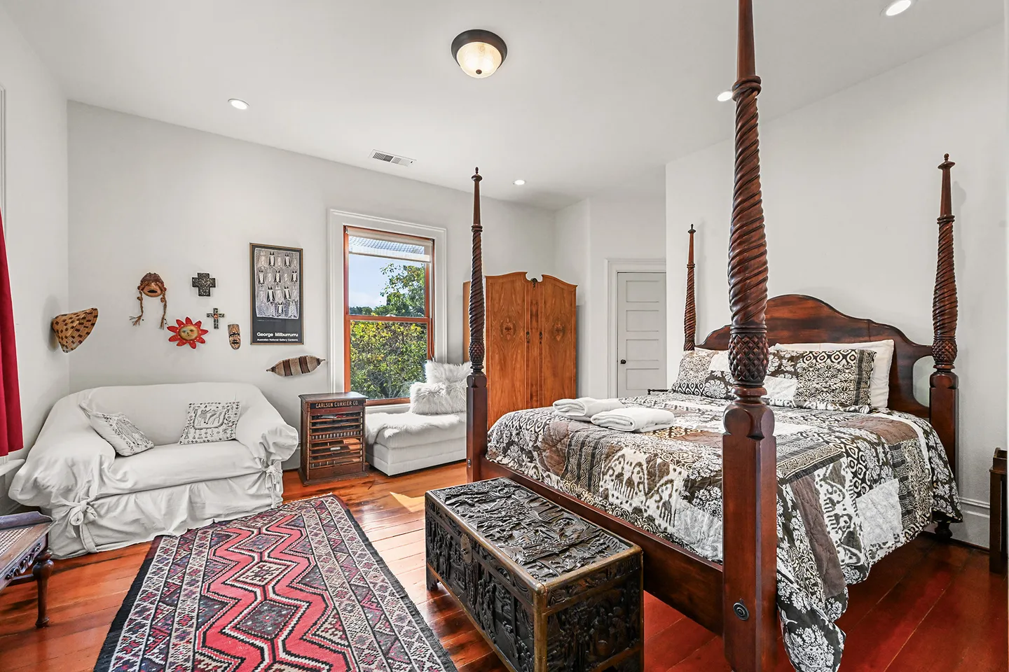 Bedroom featuring a carved four-poster wooden bed, white slipcovered sofa, pattern rug, and wood flooring.