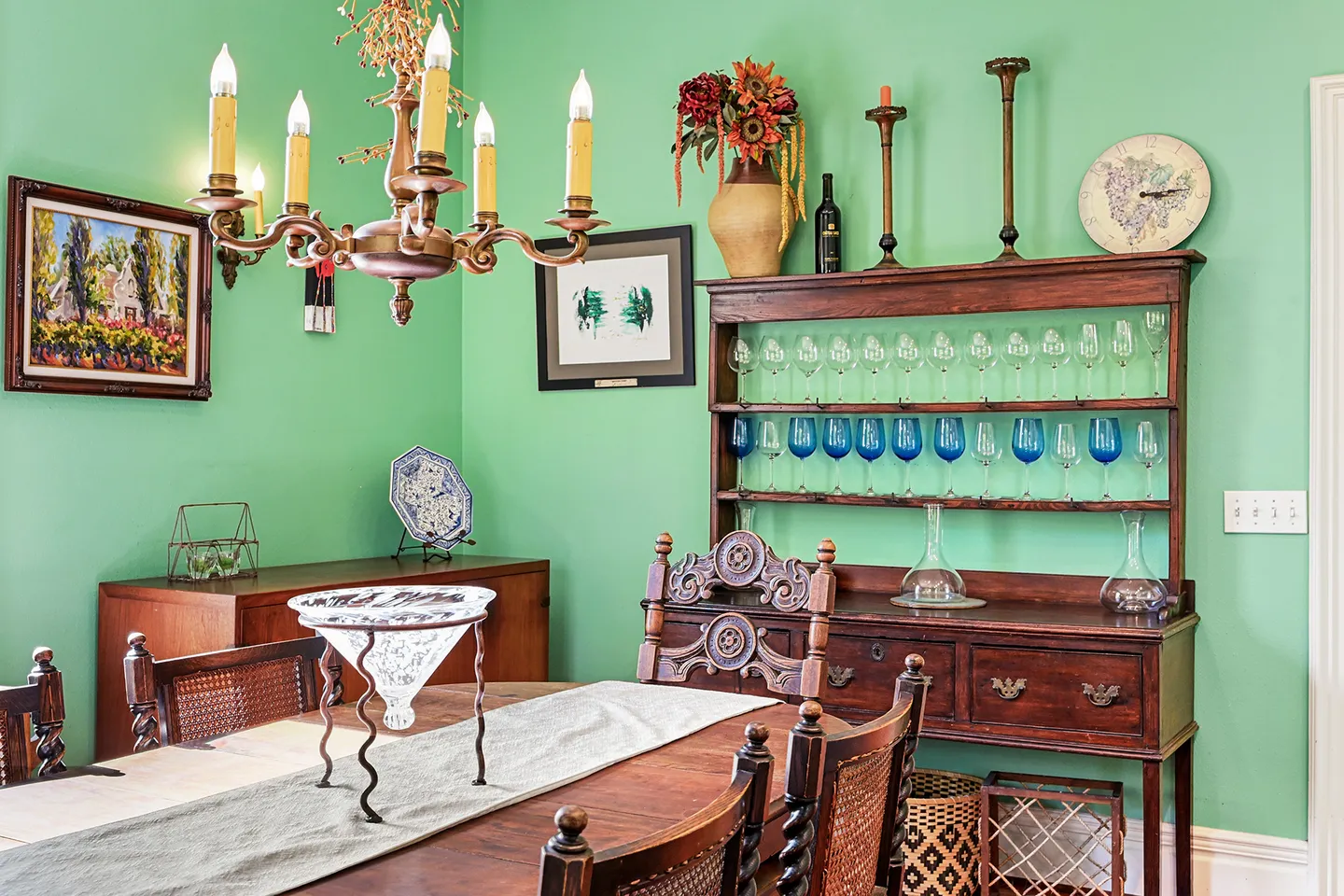 Dining room with mint green walls, a wooden dining table, a traditional wooden hutch displaying wine glasses, and a chandelier.