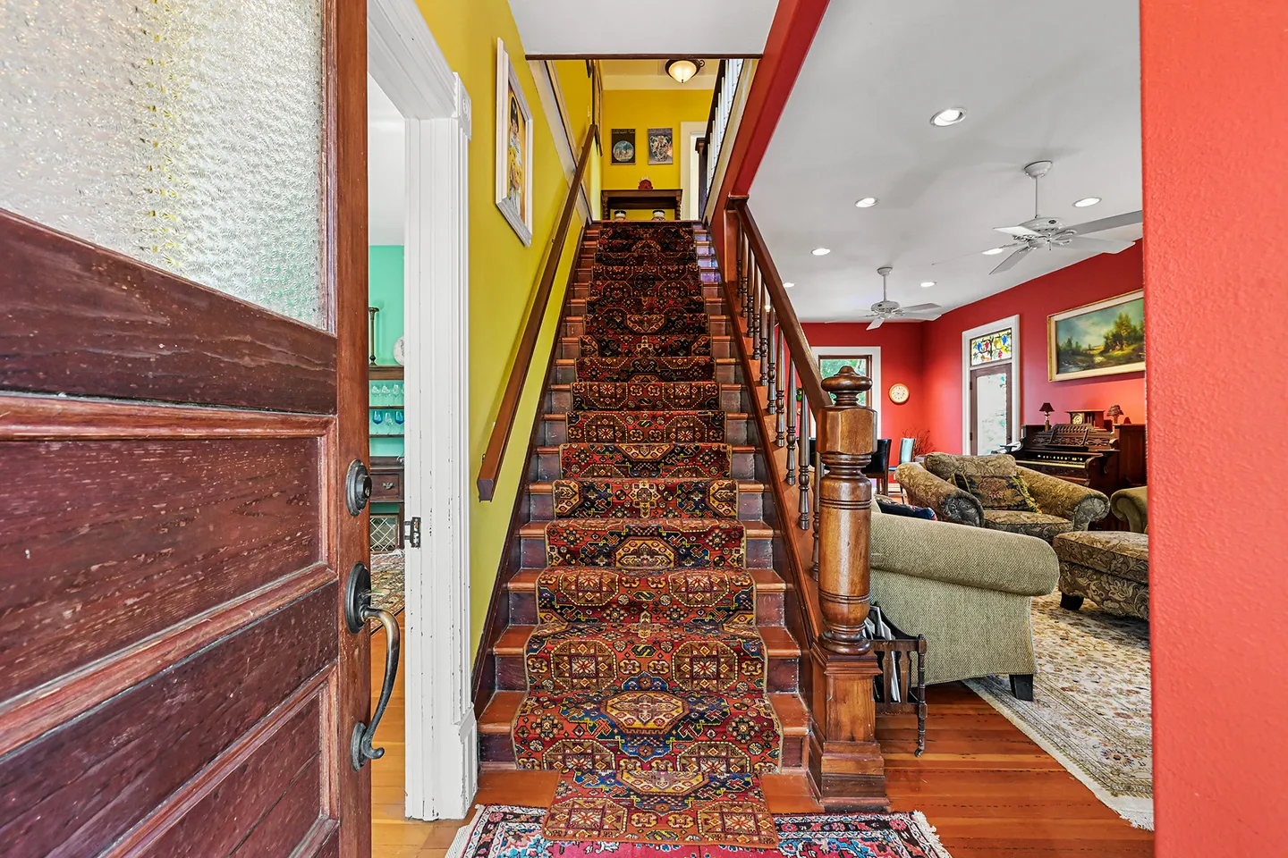 Entryway looking up a wooden staircase with an ornate patterned runner, flanked by yellow and red walls and an open living area.