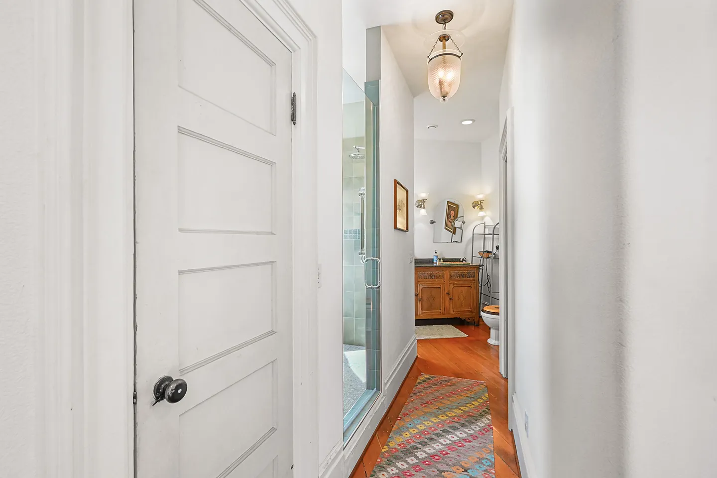 A long hallway leading into a bathroom featuring wood floors, a glass-enclosed shower, and a vanity at the far end.