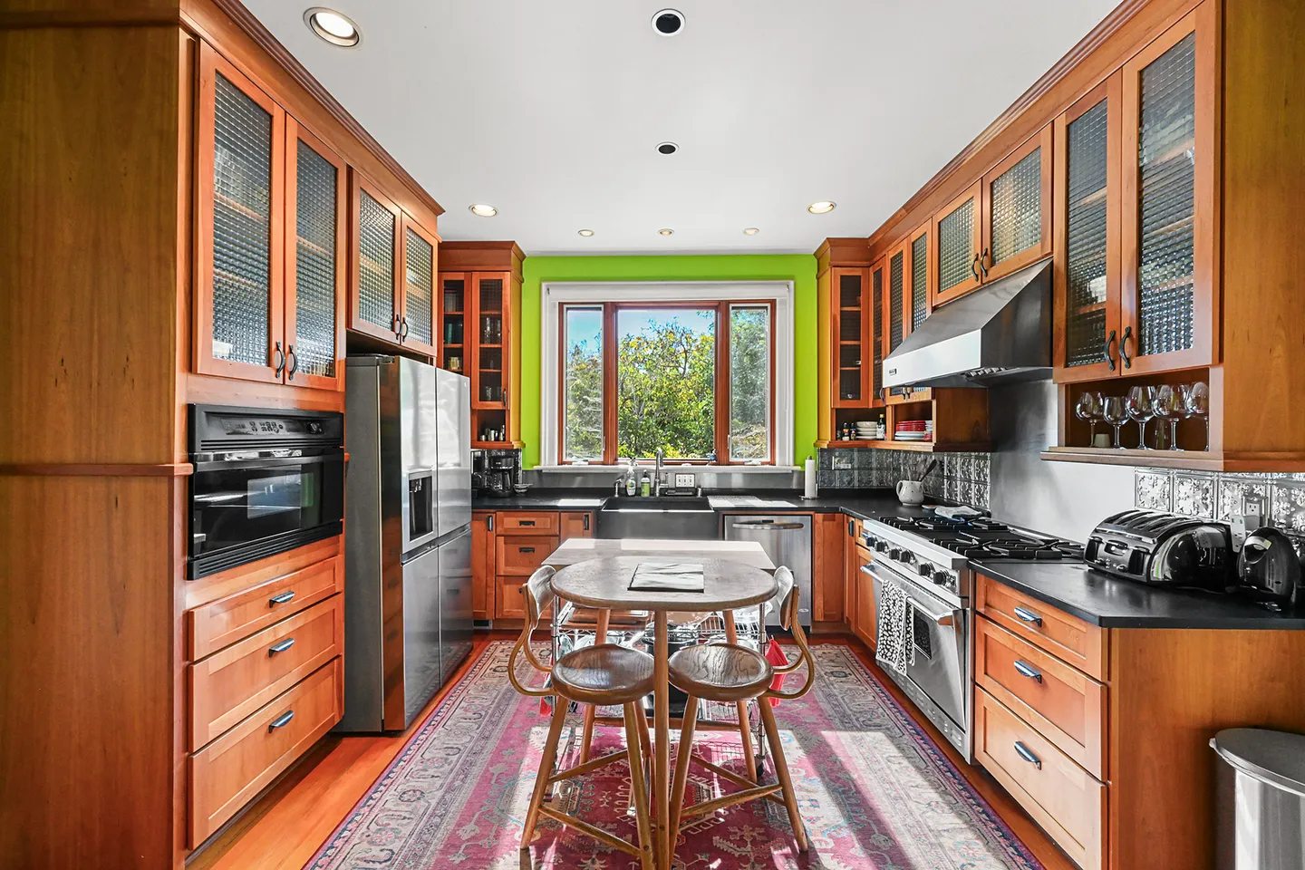 Symmetrical view of a kitchen with warm wood cabinetry, stainless steel appliances, a center island table with stools, and a bright green accent wall framing a window.