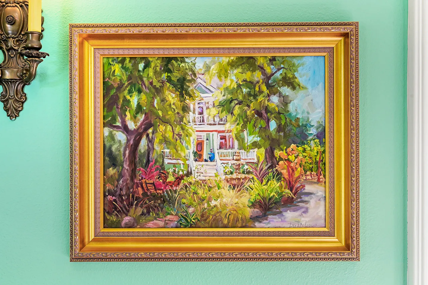 Framed painting of a historic two-story house surrounded by lush trees and garden landscaping hanging on a light green wall.