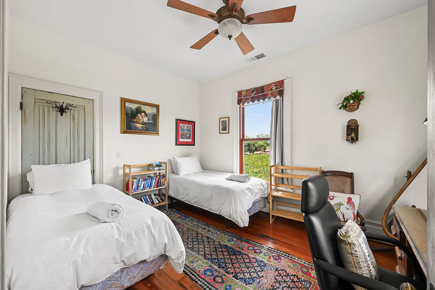 Bedroom with two twin beds dressed in white linens, wood flooring, a patterned area rug, a ceiling fan, and a desk with a black office chair.