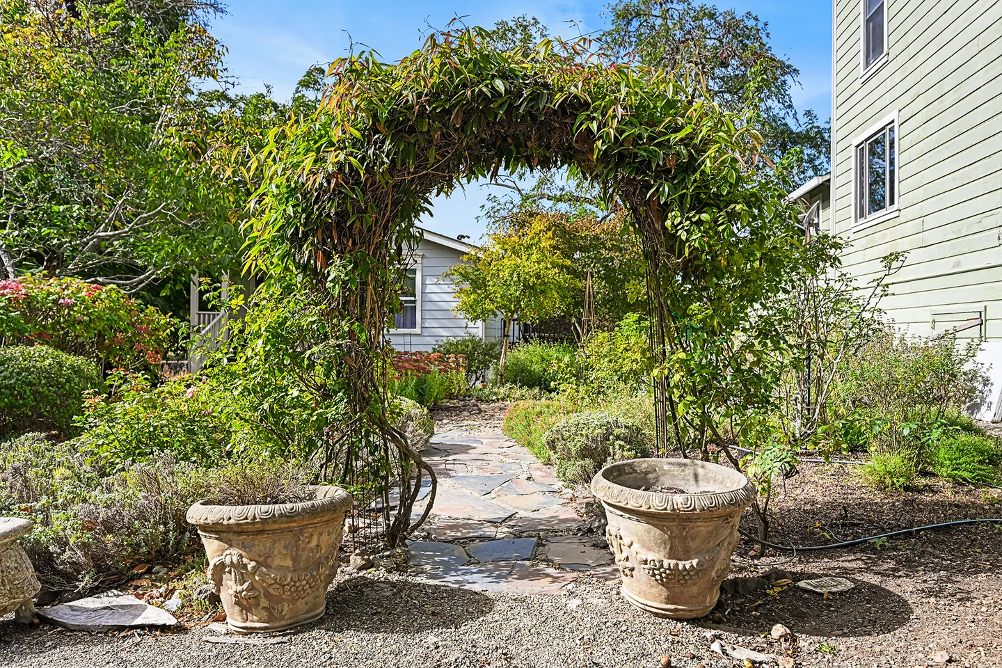 A vine-covered garden archway over a stone pathway flanked by two large ornate planters.