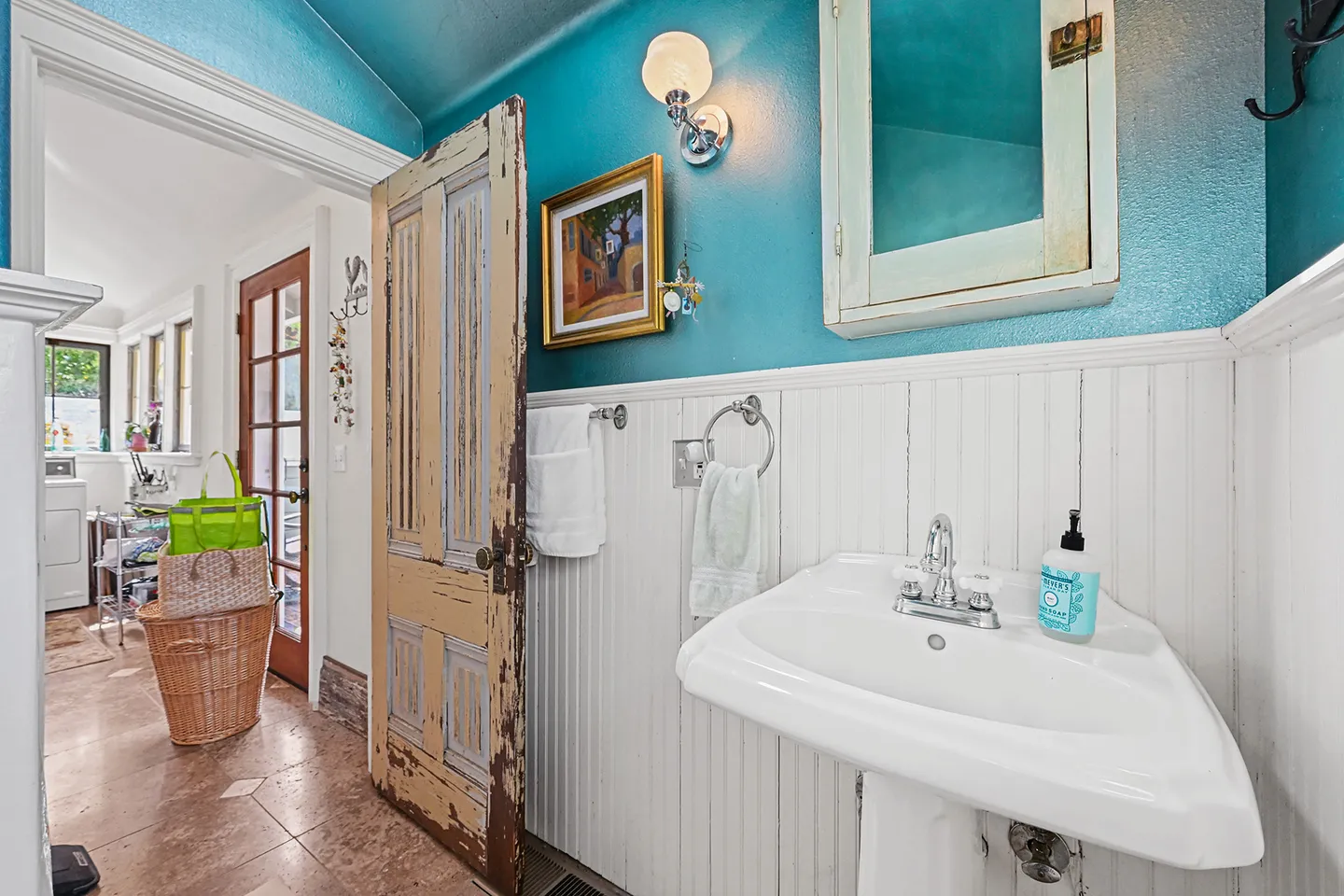 Bathroom with white pedestal sink, beadboard wainscoting, teal upper walls, and a distressed wooden door leading into an adjacent room.