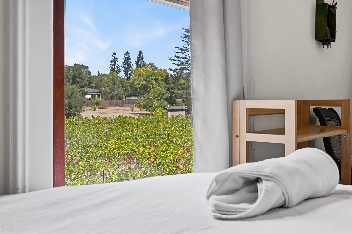 Bedroom window overlooking lush green vineyard rows and distant trees under a blue sky.