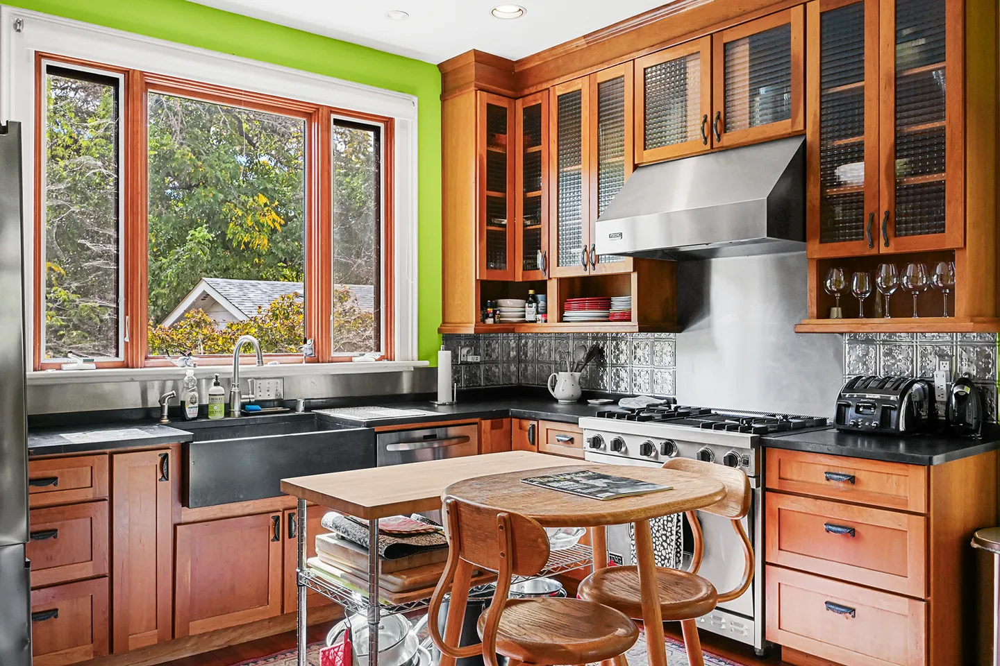 Kitchen with warm wood cabinetry, a large window overlooking trees, stainless steel gas range, and a small island with wooden chairs.