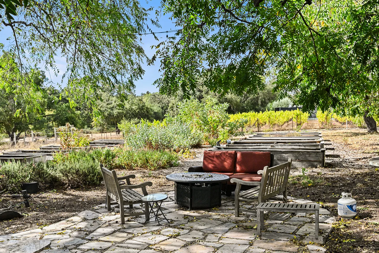Outdoor stone patio with a fire pit table, cushioned loveseat, wooden armchairs, and raised garden beds under tree shade.