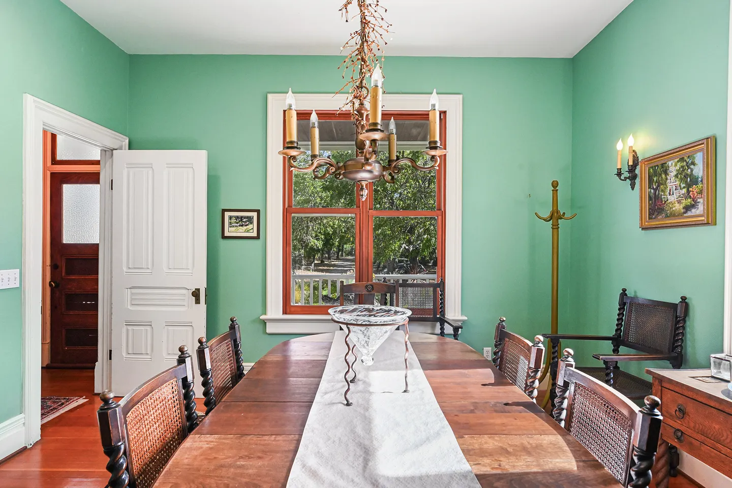 Dining room with mint green walls, a wooden dining table with chairs, an ornate chandelier, and a window overlooking greenery.