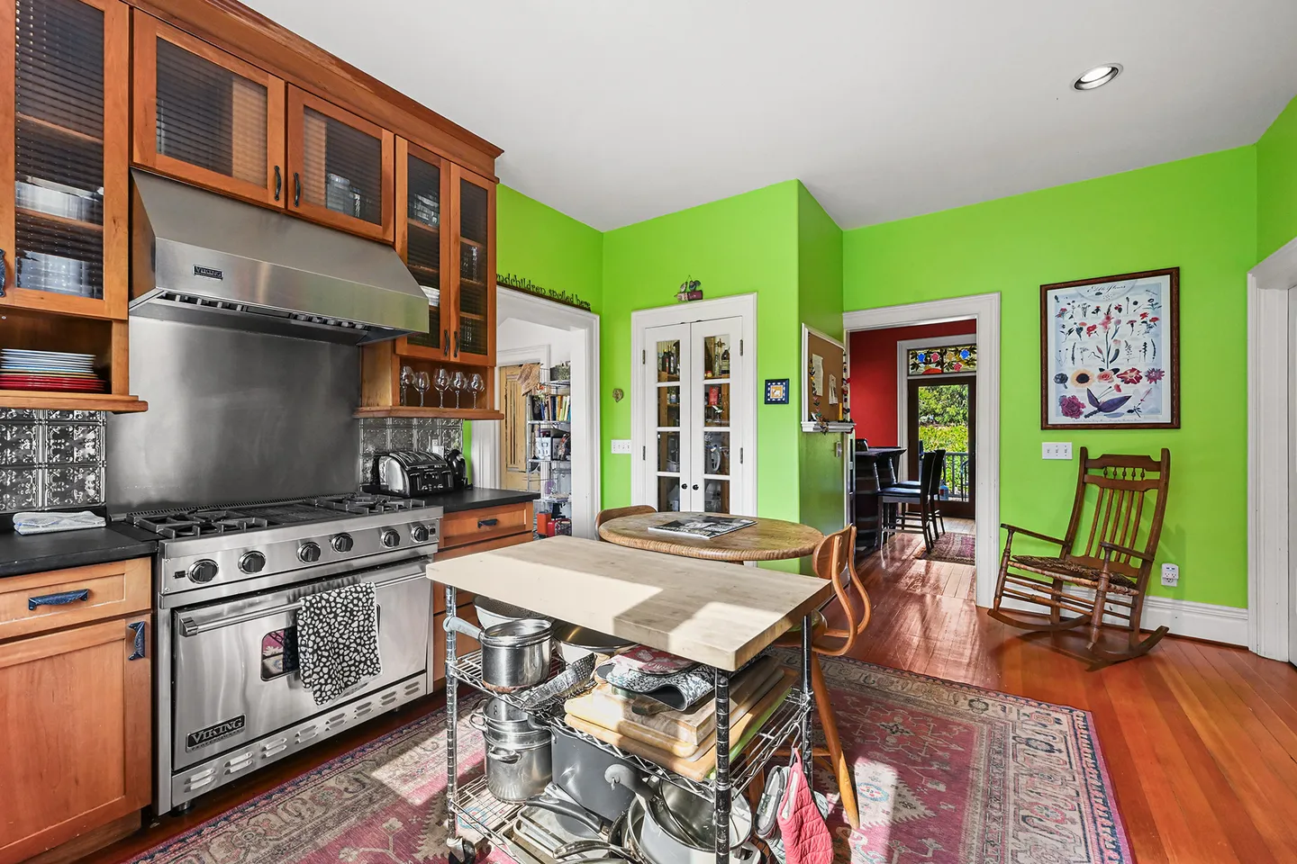 Bright kitchen with vibrant green walls, wooden cabinets, stainless steel Viking range, mobile prep island, and patterned area rug over wood floors.