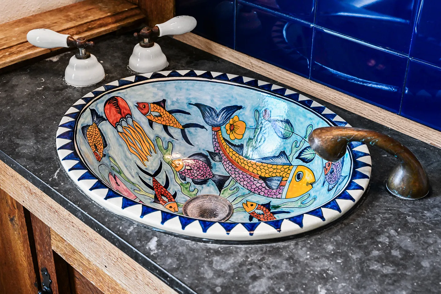 Oval bathroom sink decorated with colorful hand-painted fish motifs set into a dark speckled countertop with vintage-style handles and a curved bronze spout.