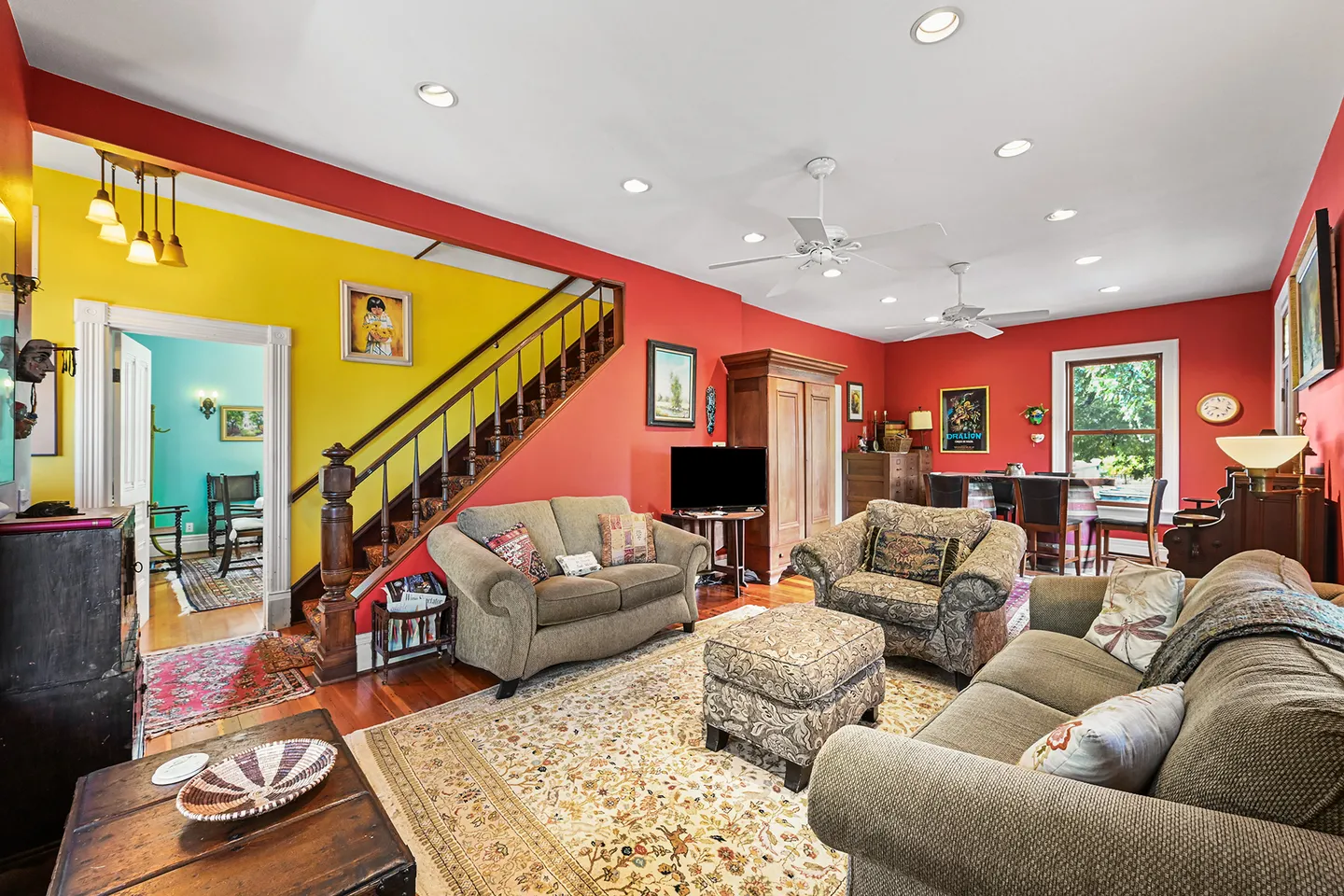 Living room with red walls, multiple sofas, patterned area rugs, a wooden staircase, and a doorway leading into a yellow hall.