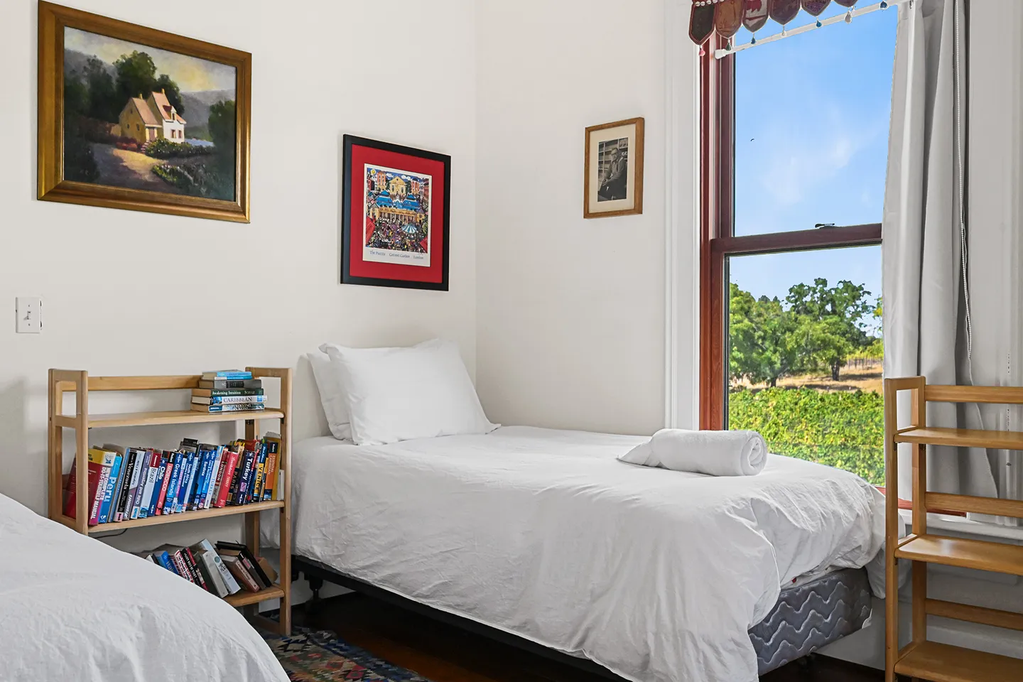 Bedroom with a single twin bed, wooden bookshelves, and a tall window looking out at trees under a blue sky.