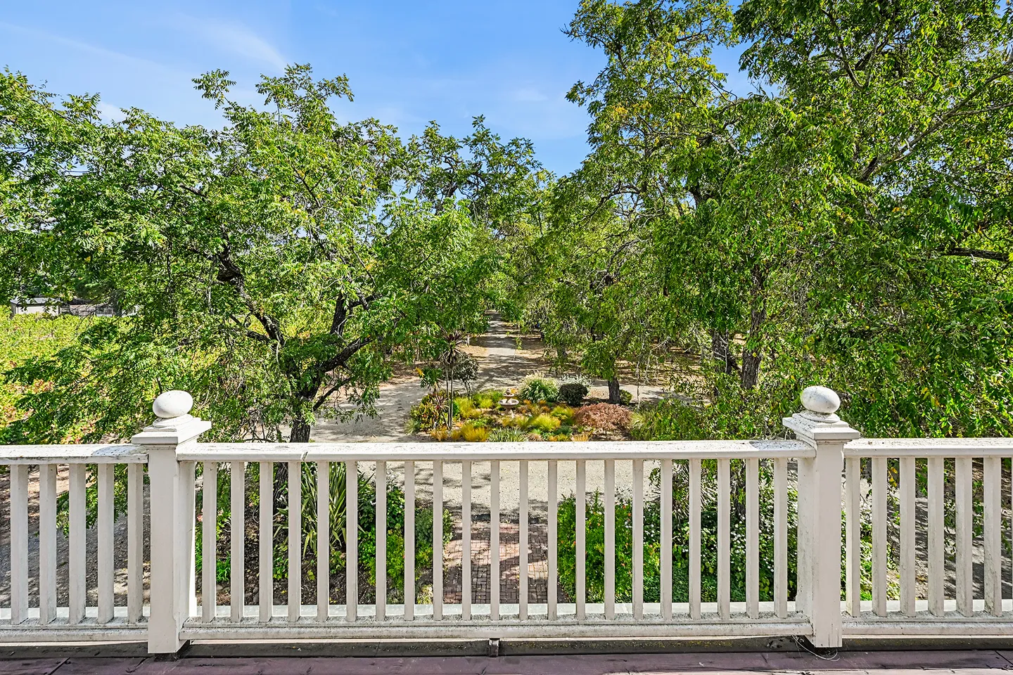 View from an elevated balcony with a white railing looking out over a garden path and mature green trees under a clear blue sky.