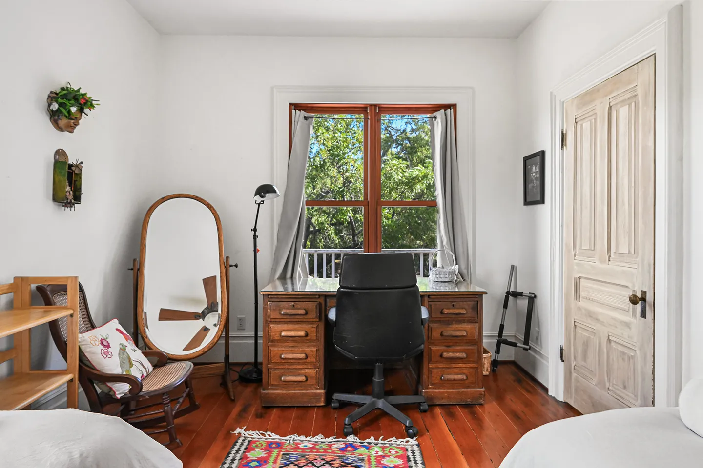 Bedroom work nook with a wooden desk, office chair, large standing mirror, rocking chair, and wood-framed window looking out to greenery.