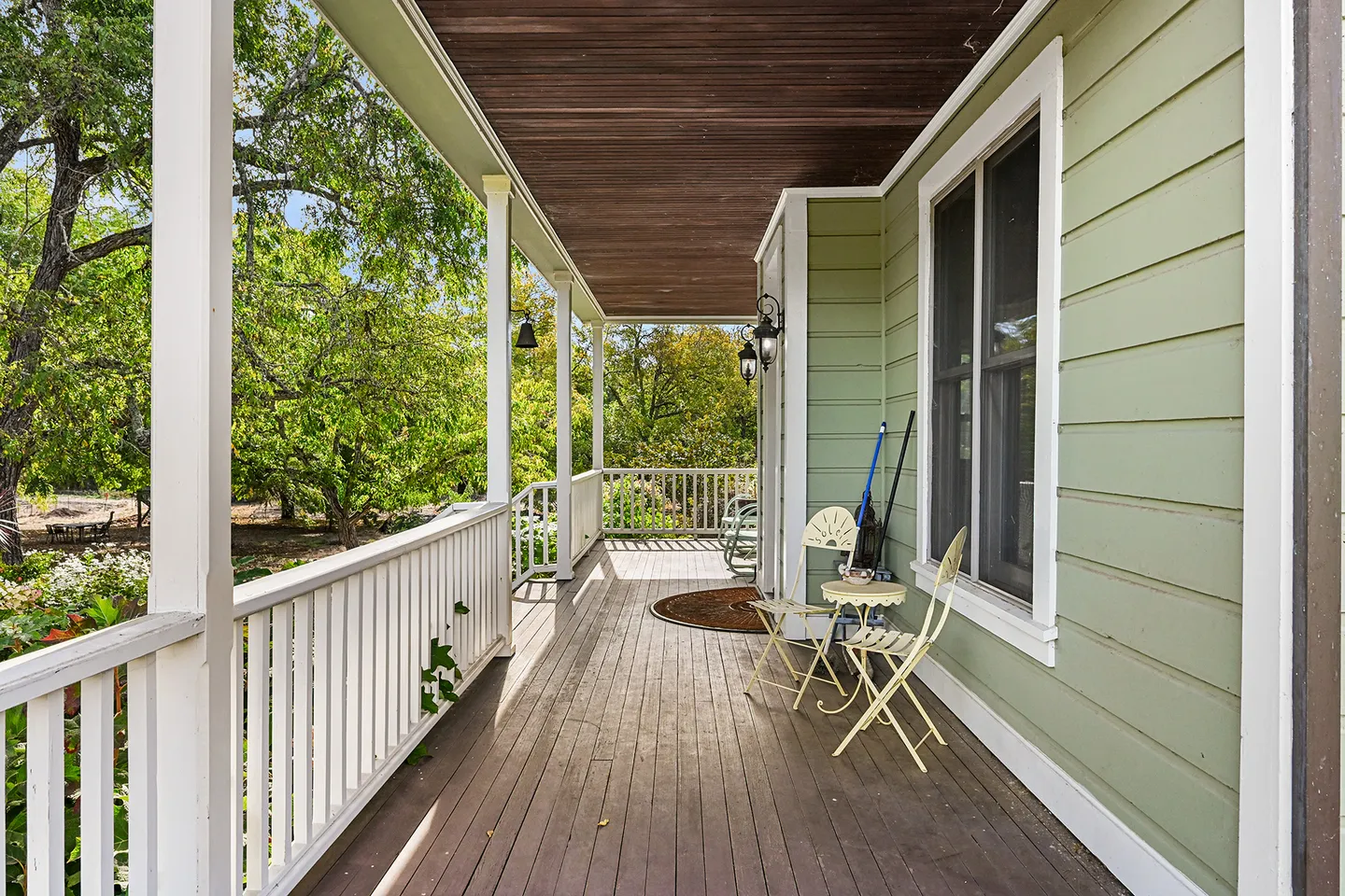 A long covered porch with dark wood floor planks, white railings, and light green siding overlooking surrounding trees.