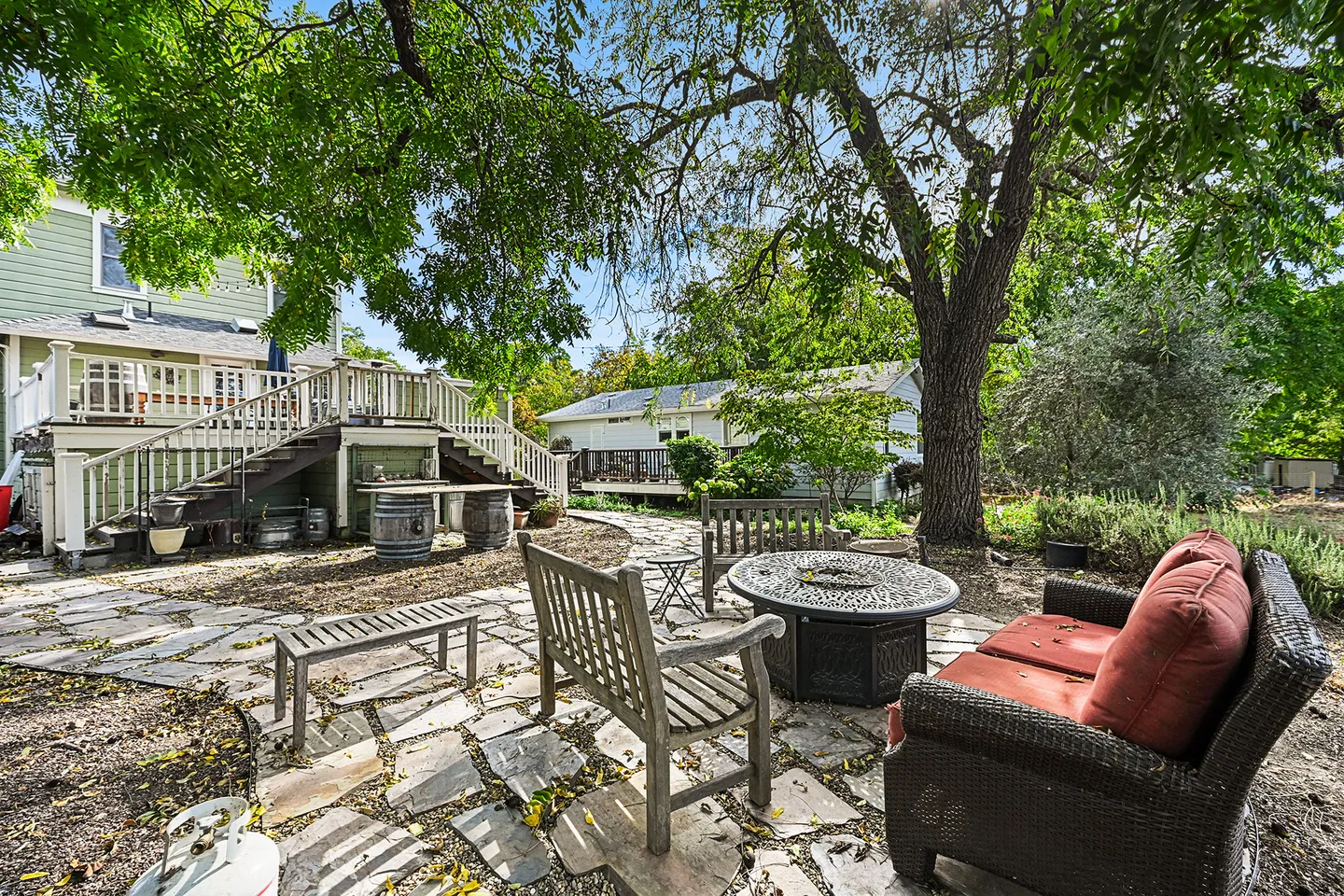 A backyard stone patio featuring a fire pit, wooden benches, an outdoor sofa, and stairs leading to an elevated deck.