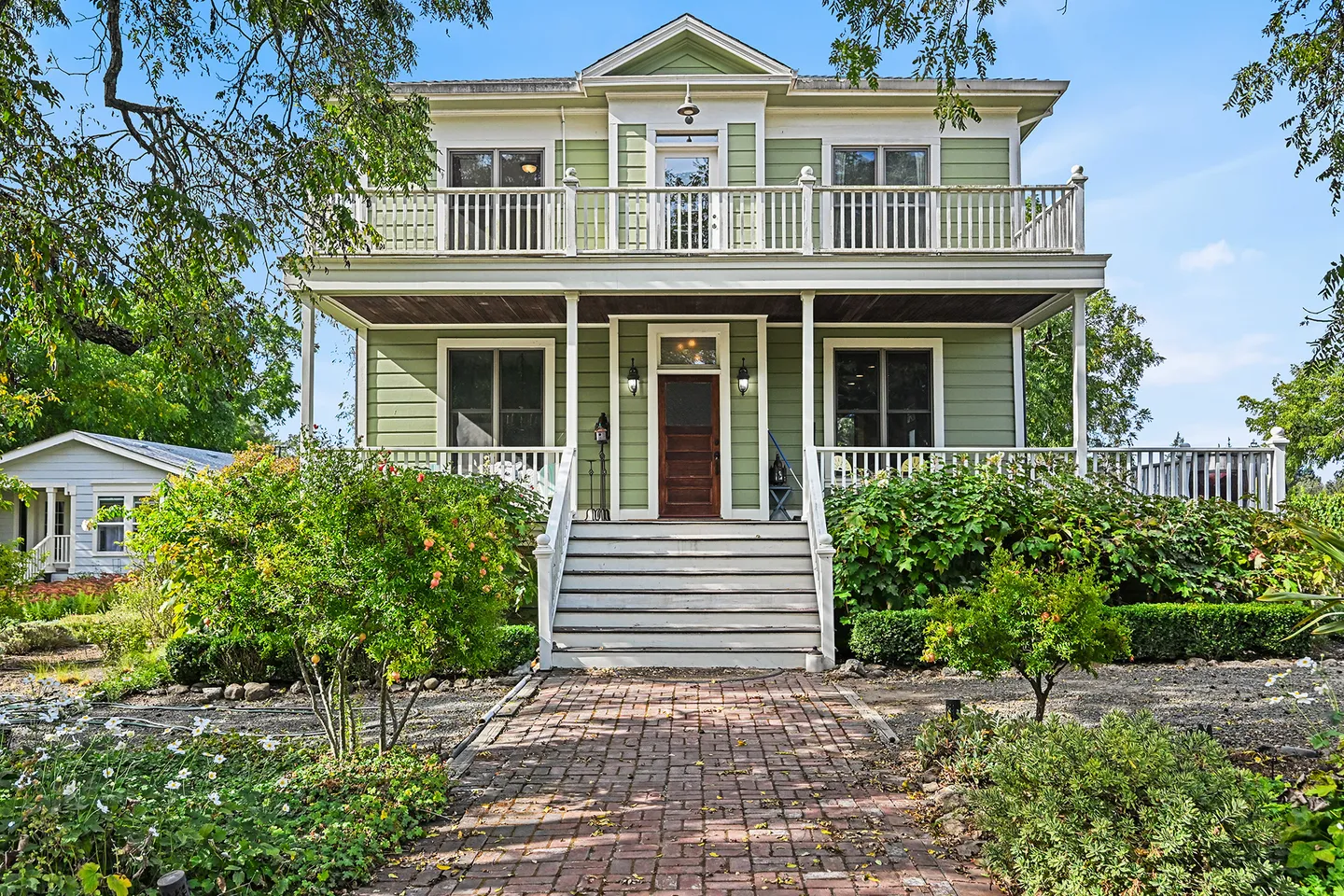 Exterior front view of a two-story green house with white trim, a covered front porch, upper balcony, and brick walkway.