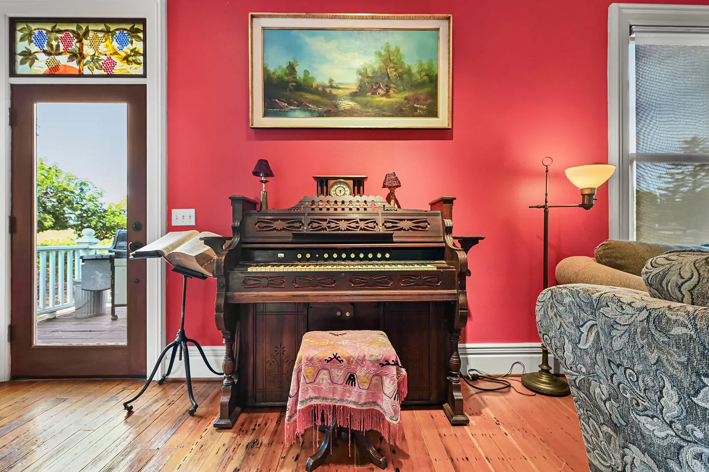 Antique pump organ with a stool against a vibrant coral-red wall, alongside wood flooring, a stained glass door transom, and an oil painting.