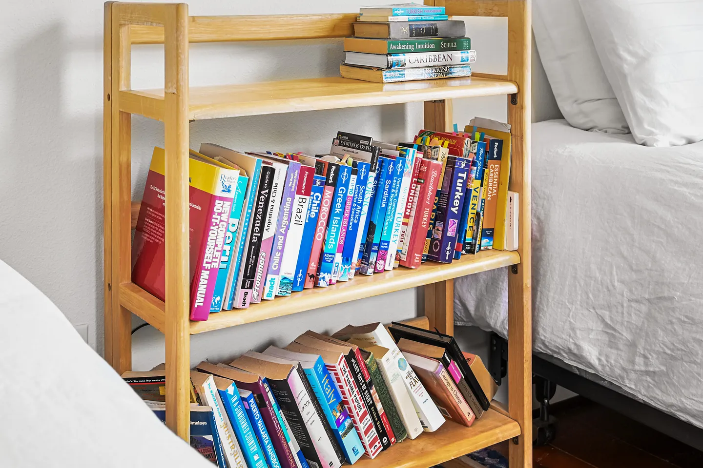 A light wood bookshelf filled with assorted travel guidebooks and other books between two beds.