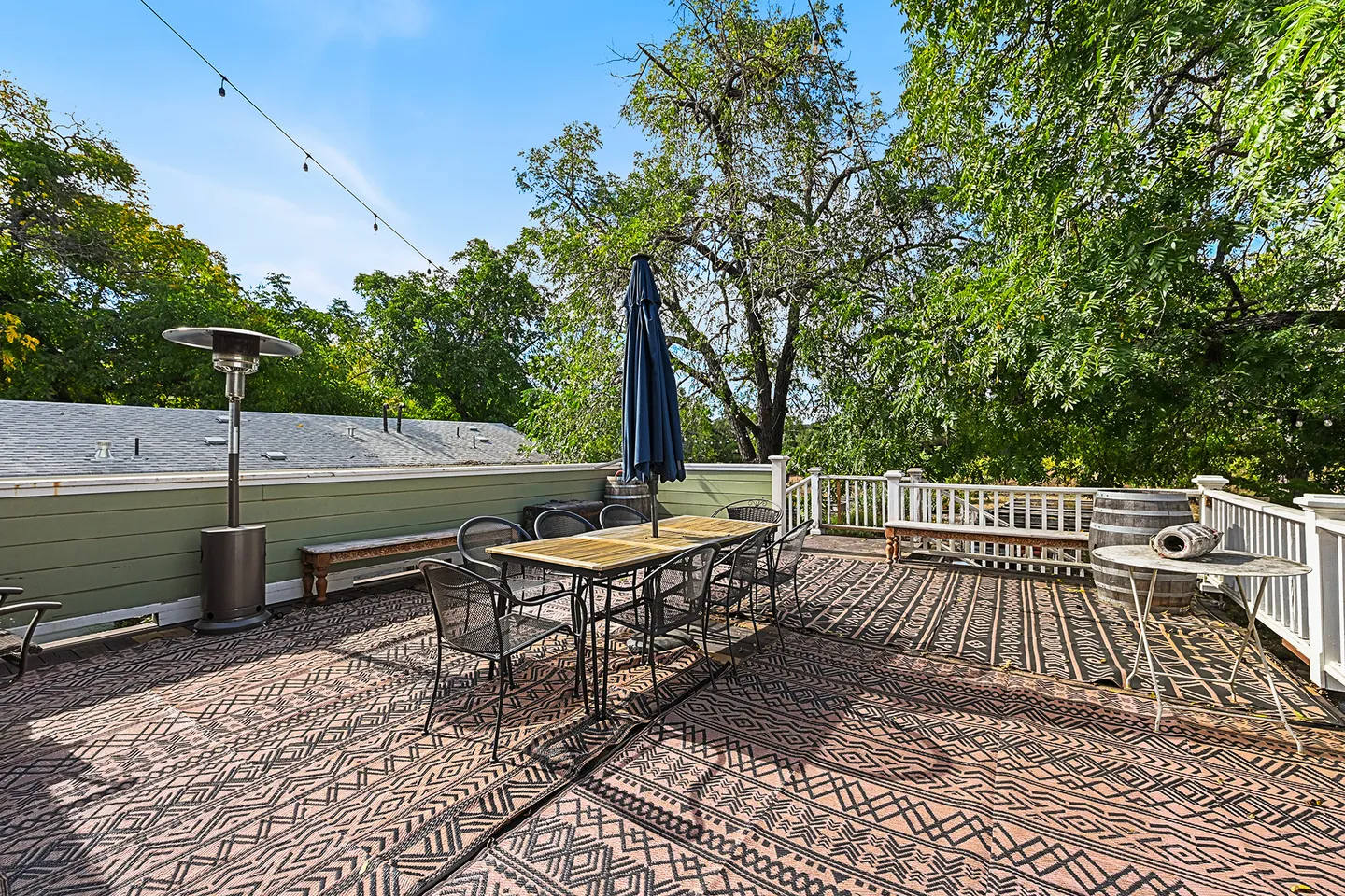 Elevated outdoor patio with geometric outdoor rugs, a dining table with chairs and an umbrella, a patio heater, and surrounding mature trees.