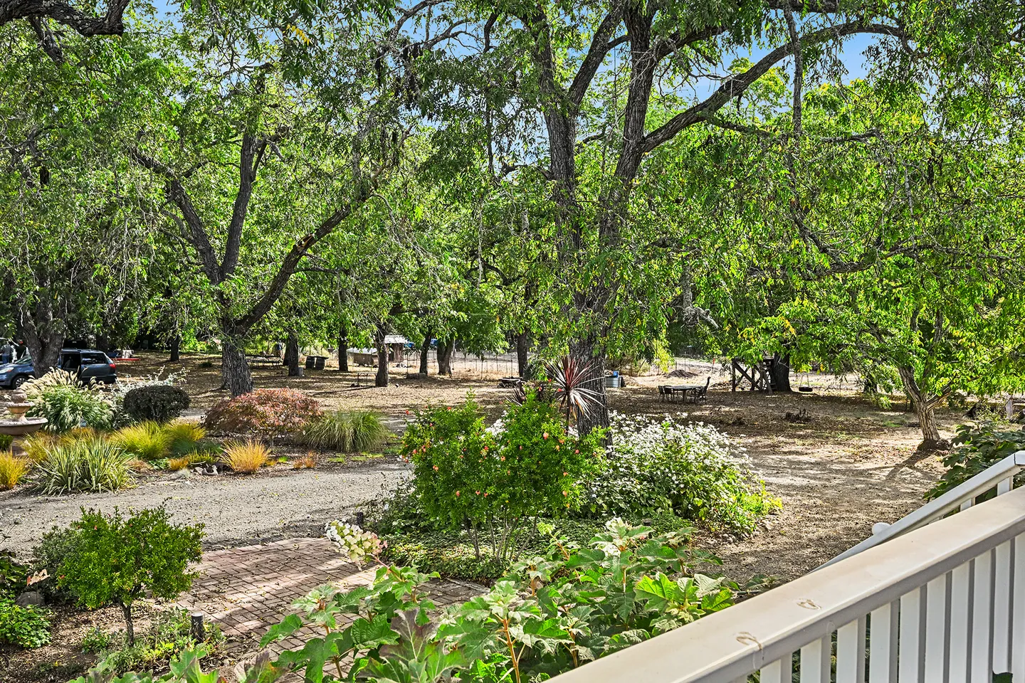 Elevated view from a porch railing overlooking a spacious yard shaded by large trees with garden beds and a brick pathway.