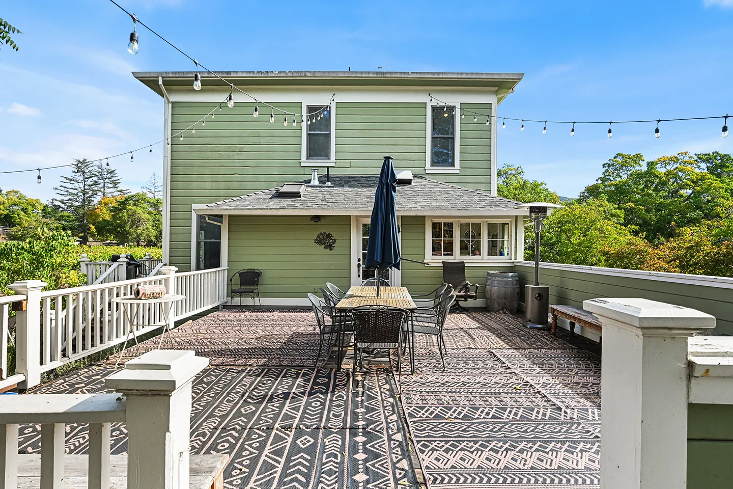 Spacious outdoor deck with dining table, patterned rugs, and string lights outside a green two-story house.