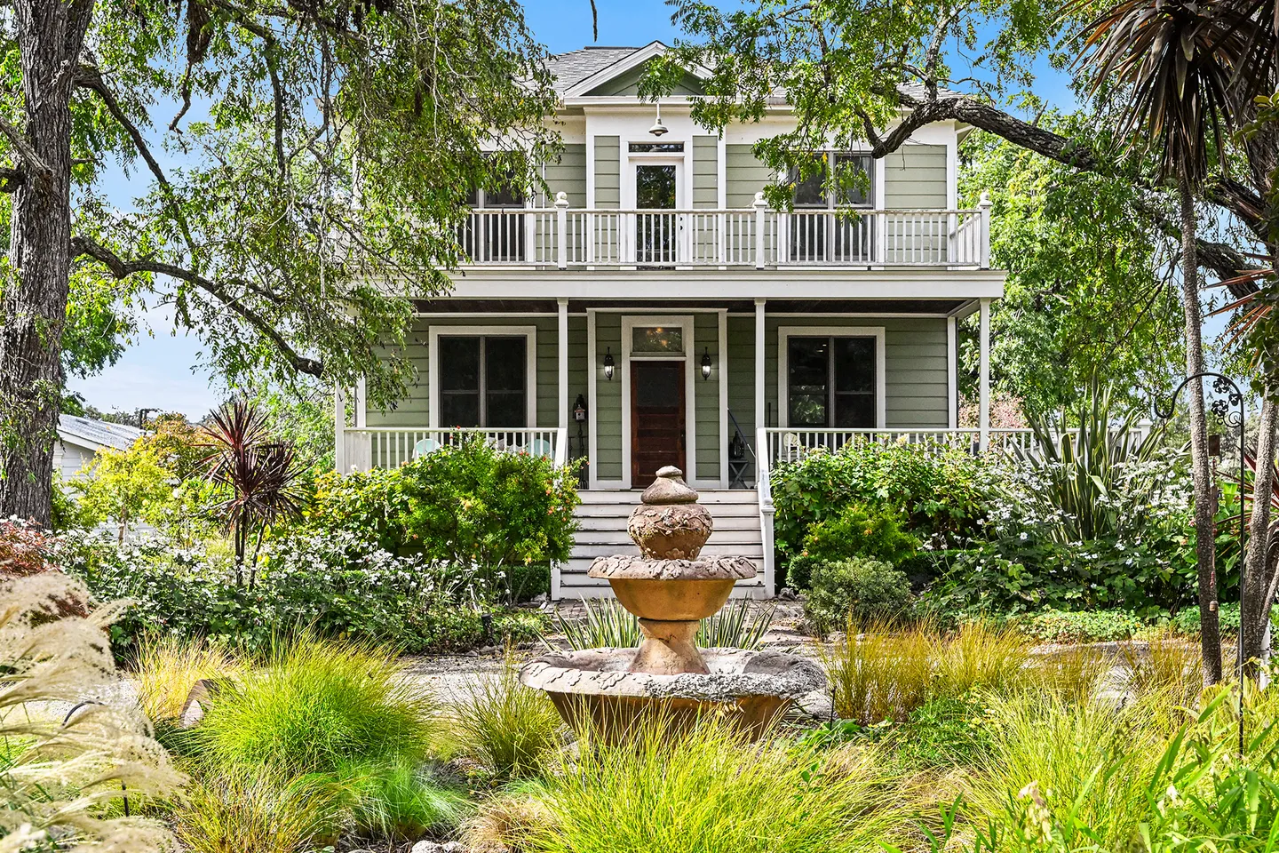Front view of a two-story green house with white trim, upper and lower porches, and a stone fountain in the garden.