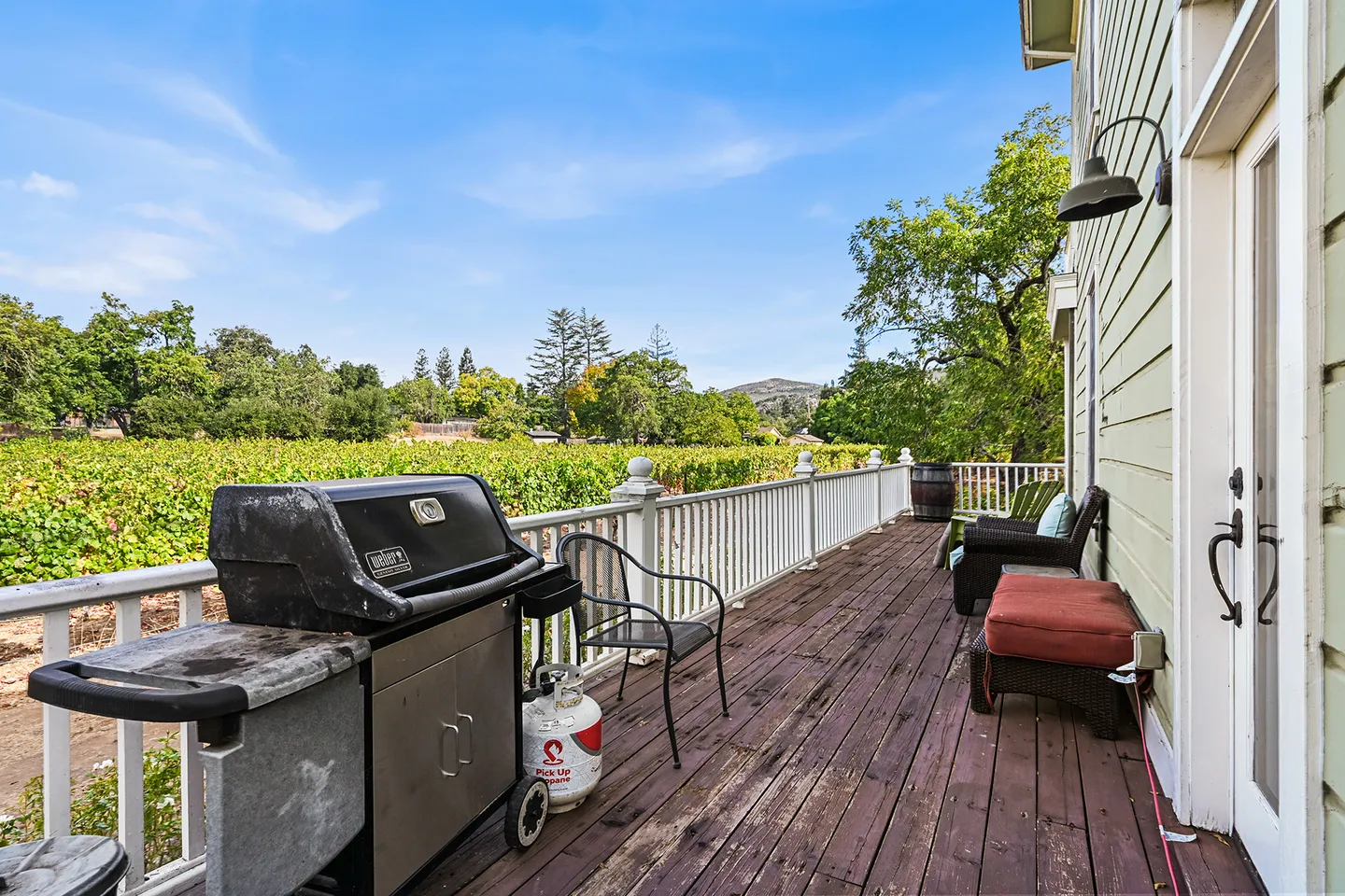 Wooden deck with a barbecue grill, railing, and seating overlooking a lush vineyard under a clear blue sky.