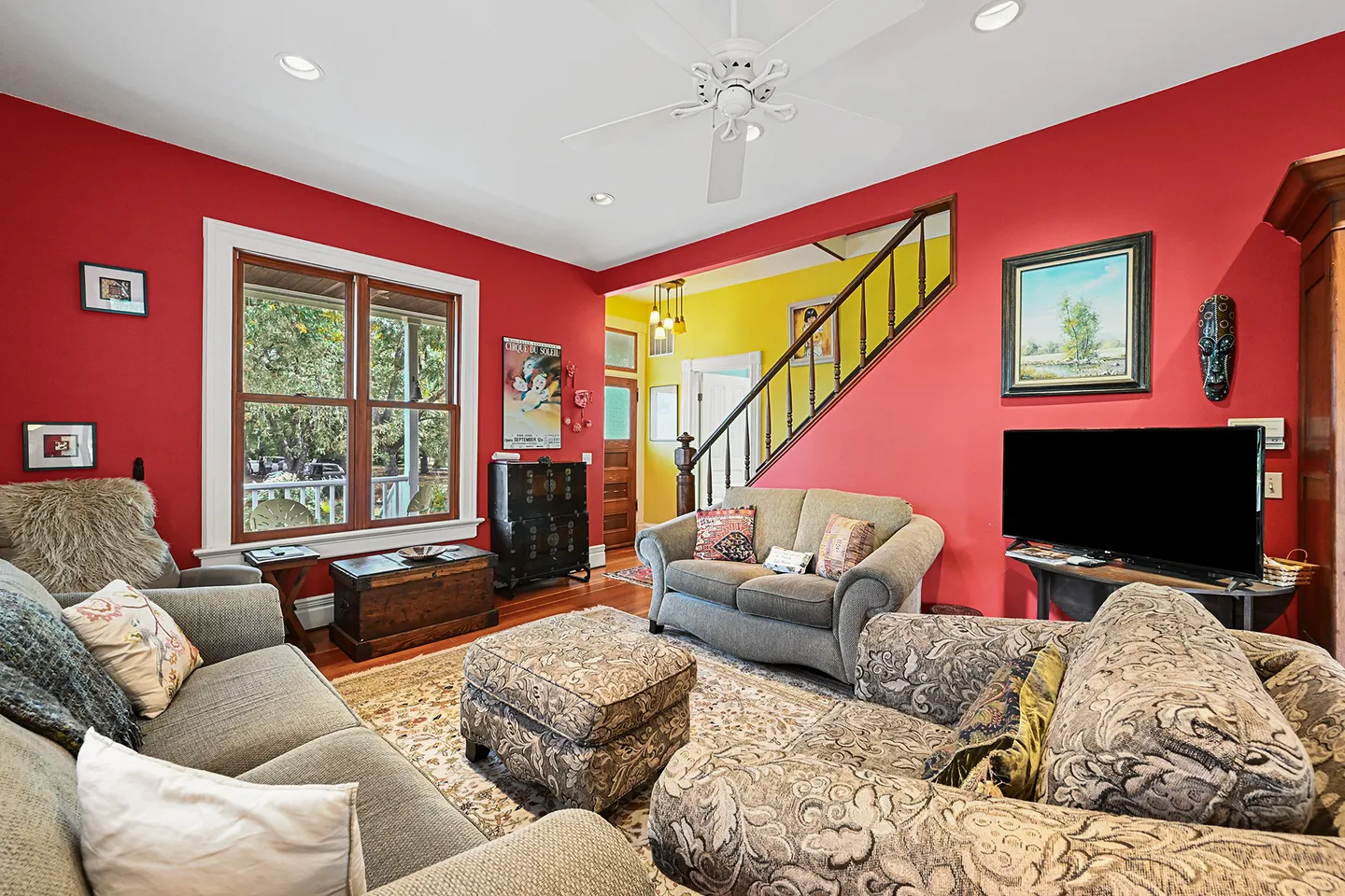 Living room with red walls, upholstered sofas, an ottoman, wood floors, a flat-screen television, and a staircase leading upstairs.