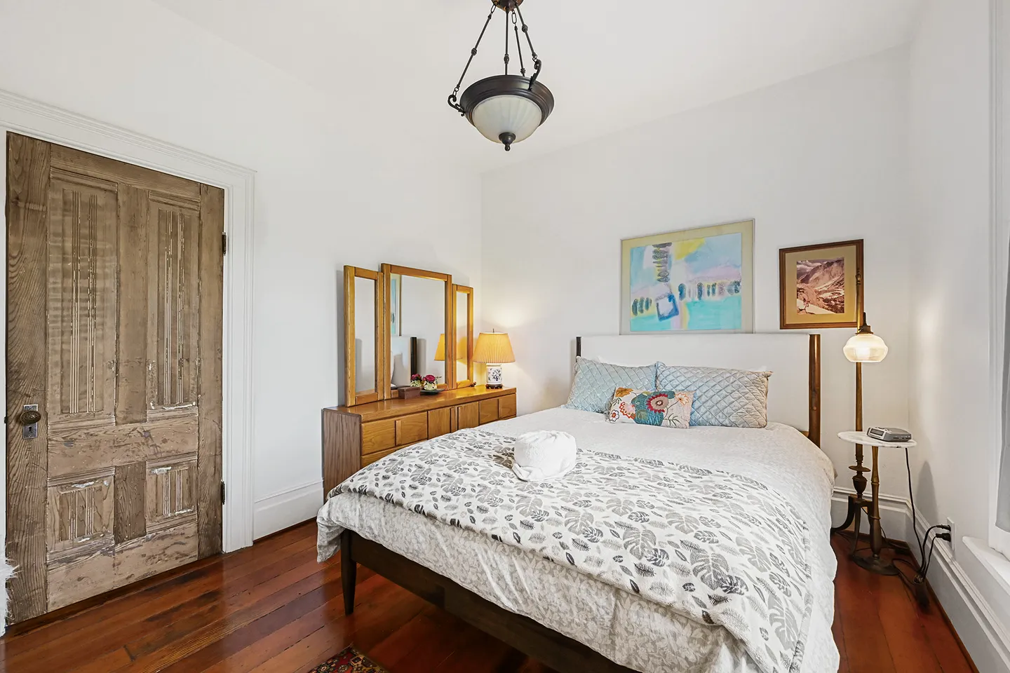 Bedroom with wooden floors, a rustic wood door, dresser with mirror, and a made bed under a hanging light fixture.