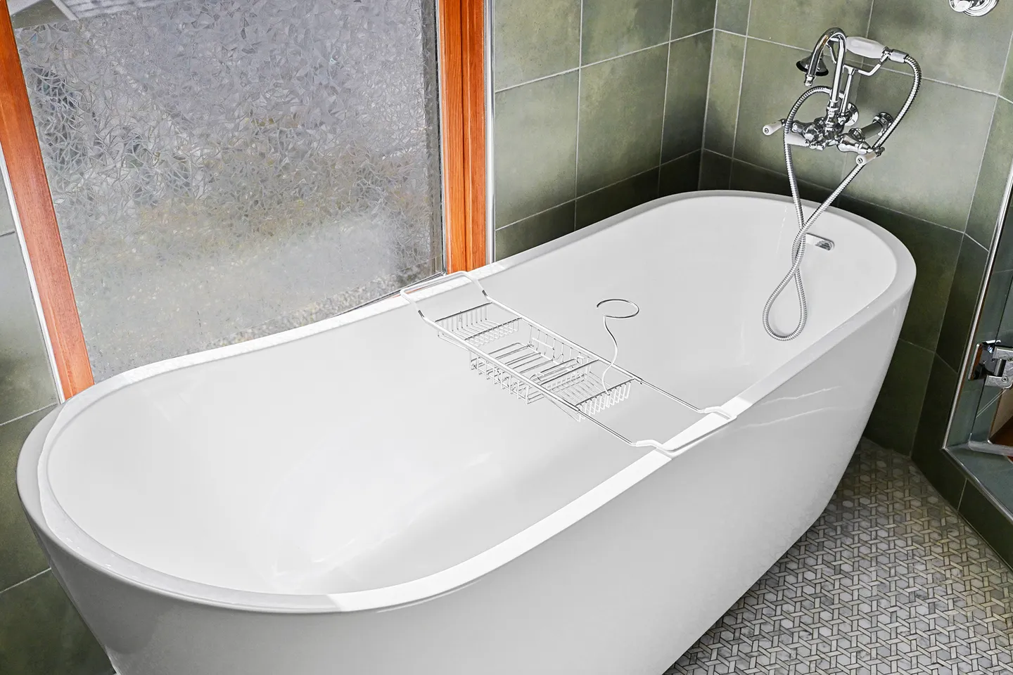 A freestanding white soaking bathtub fitted with a metal bath caddy, situated next to a frosted window and green wall tiles.
