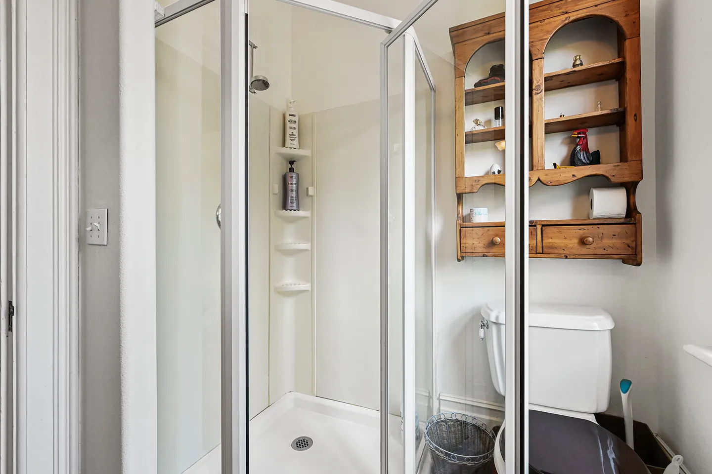 Bathroom showing a glass-enclosed corner shower next to a toilet and a wooden wall-mounted shelving unit.