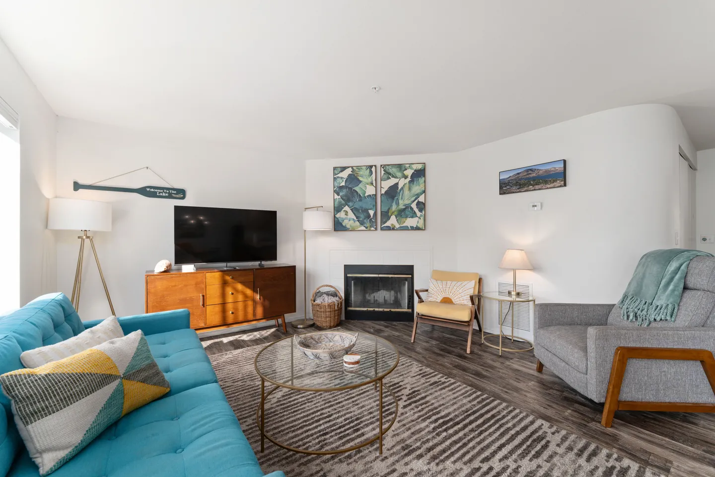 Bright living room featuring a turquoise couch, a fireplace, television on a wooden console, and varied accent chairs around a round glass coffee table.