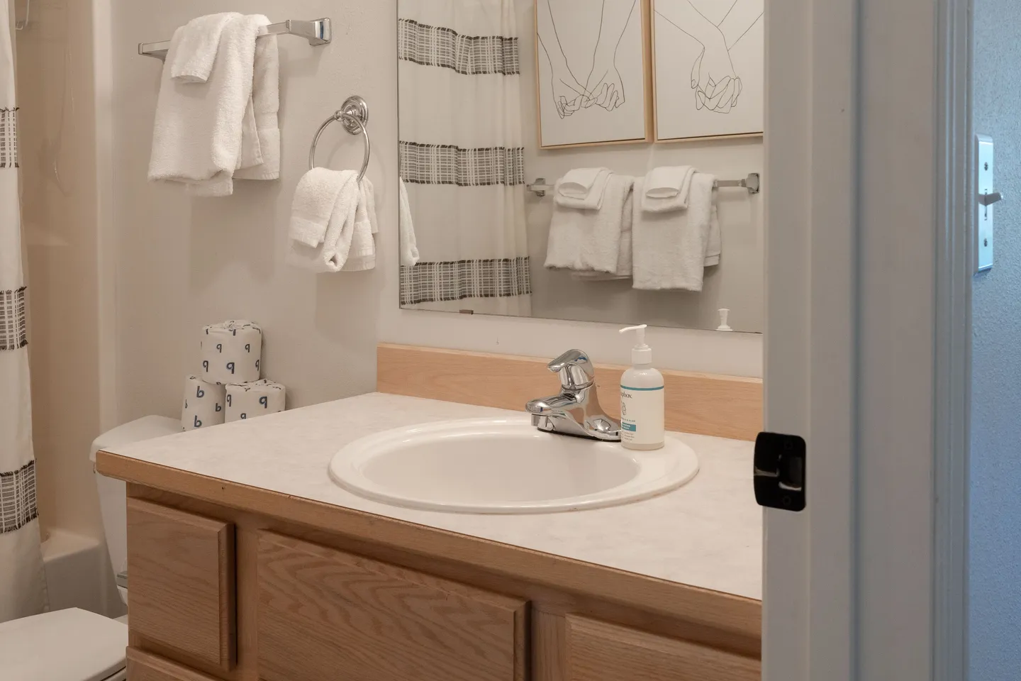 Bathroom vanity with a light countertop, wood cabinets, mirror reflecting towels and artwork, and a partial view of the toilet.