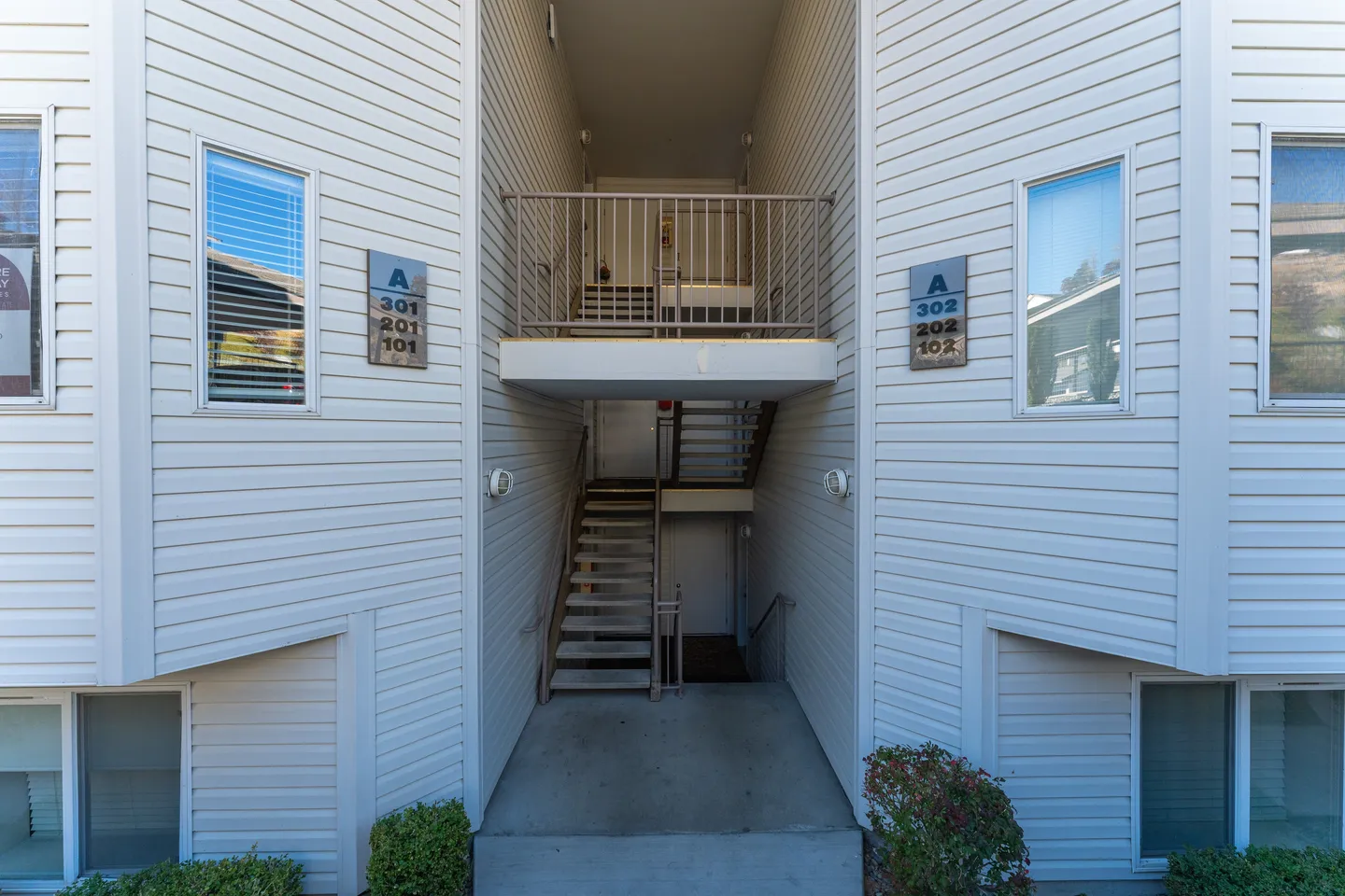 Exterior entryway and central open-air stairwell of an apartment building with white horizontal siding and unit signage.