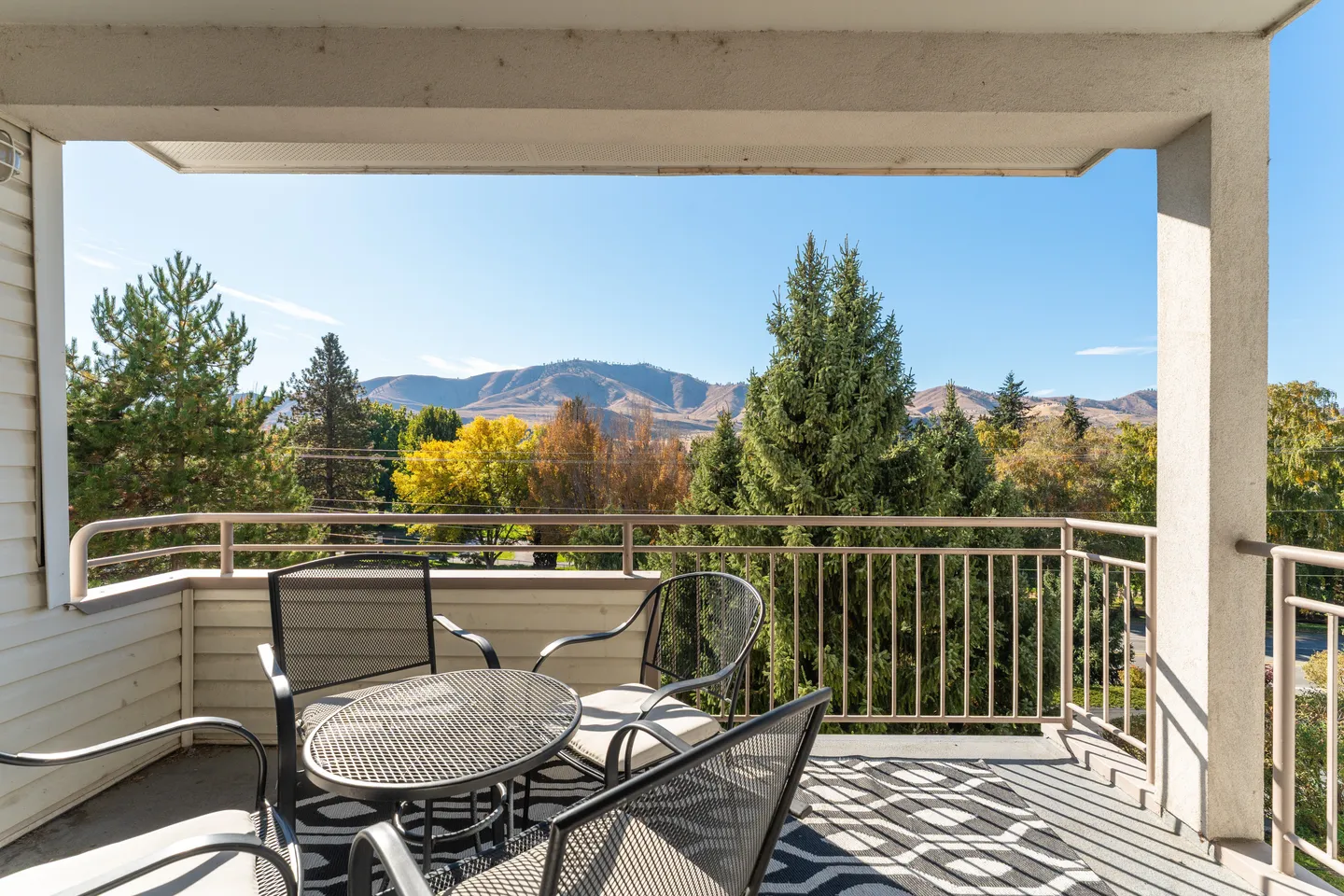 A covered balcony with outdoor chairs, a small table, and a geometric rug overlooking trees and distant mountains under a blue sky.