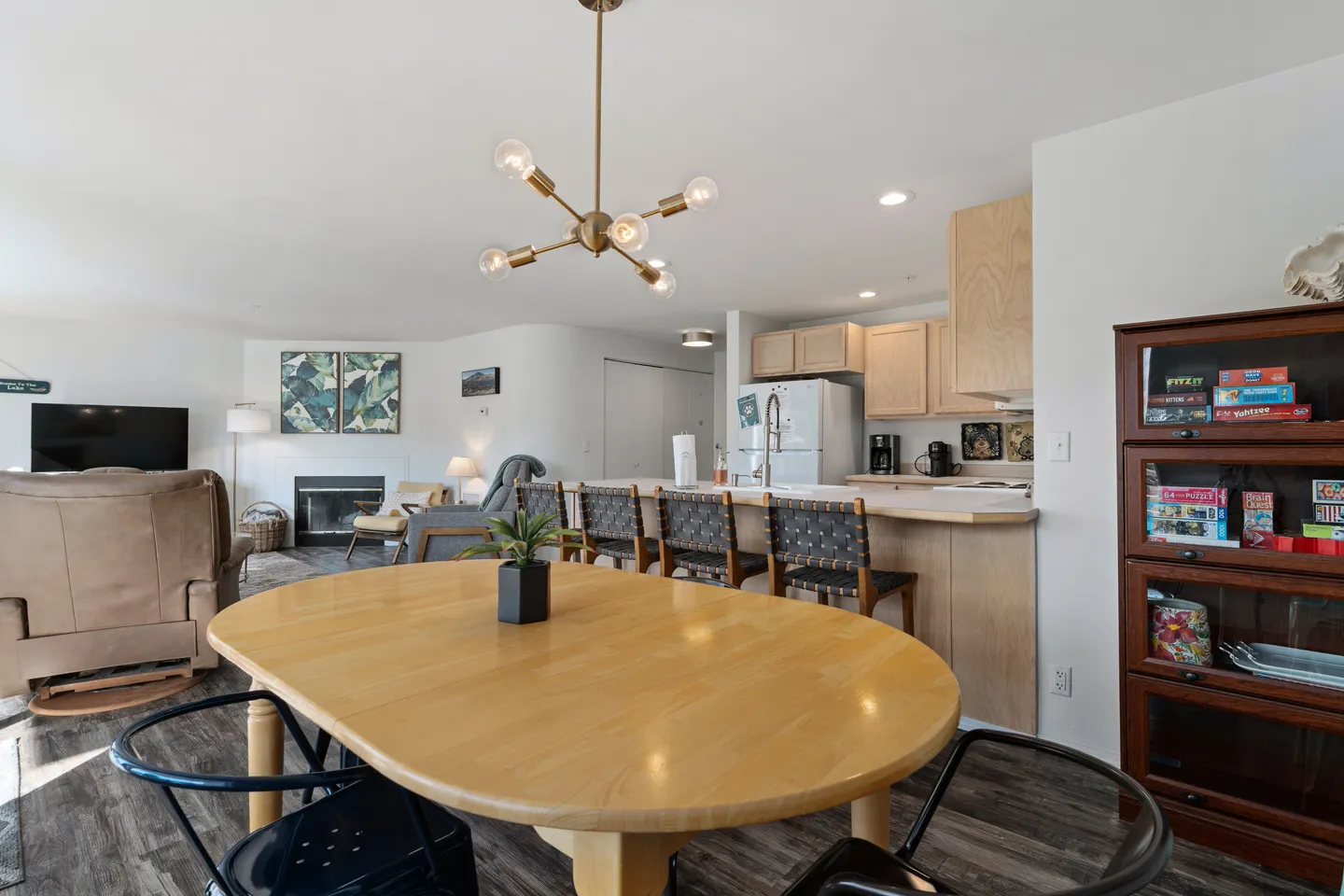 Open-concept living space with an oval dining table, kitchen breakfast bar with stools, and a living area with a fireplace in the background.