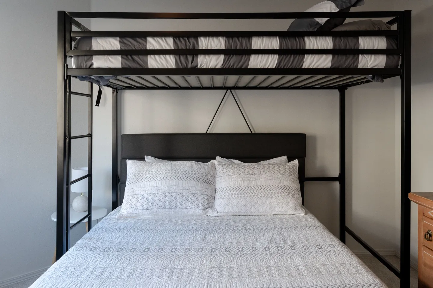 A black metal loft bed frame over a double bed with an upholstered headboard, dressed in white patterned bedding.