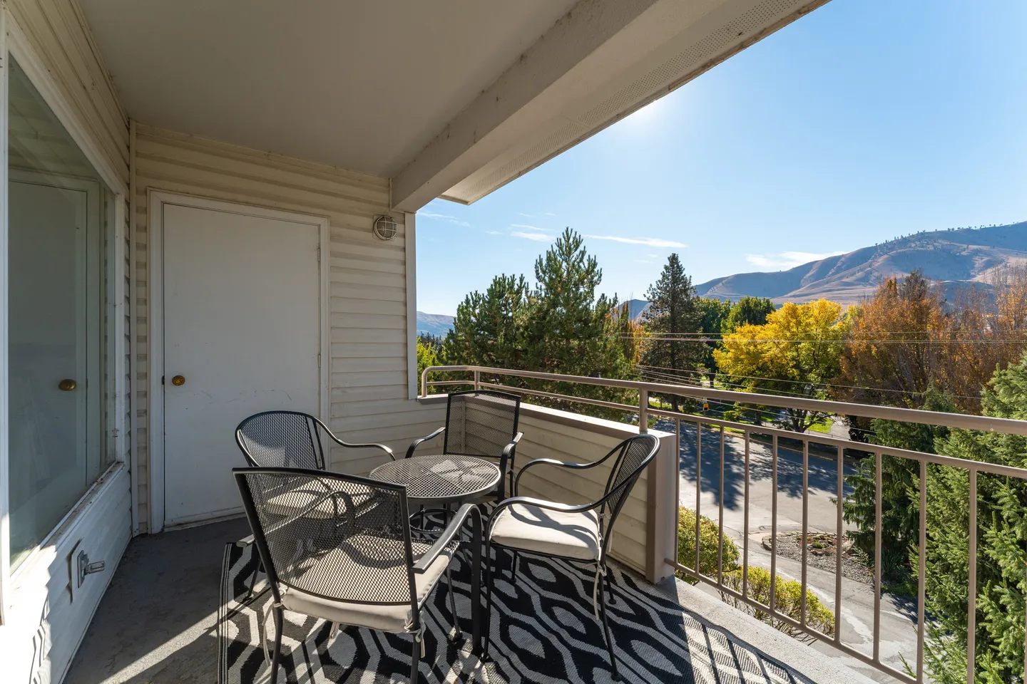 Balcony with patio chairs and a small round table overlooking trees, a street, and distant hills under a blue sky.