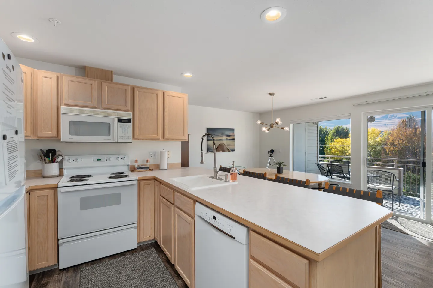Open-concept kitchen with light wood cabinetry, white appliances, and an island facing a dining area and sliding glass doors to a balcony.