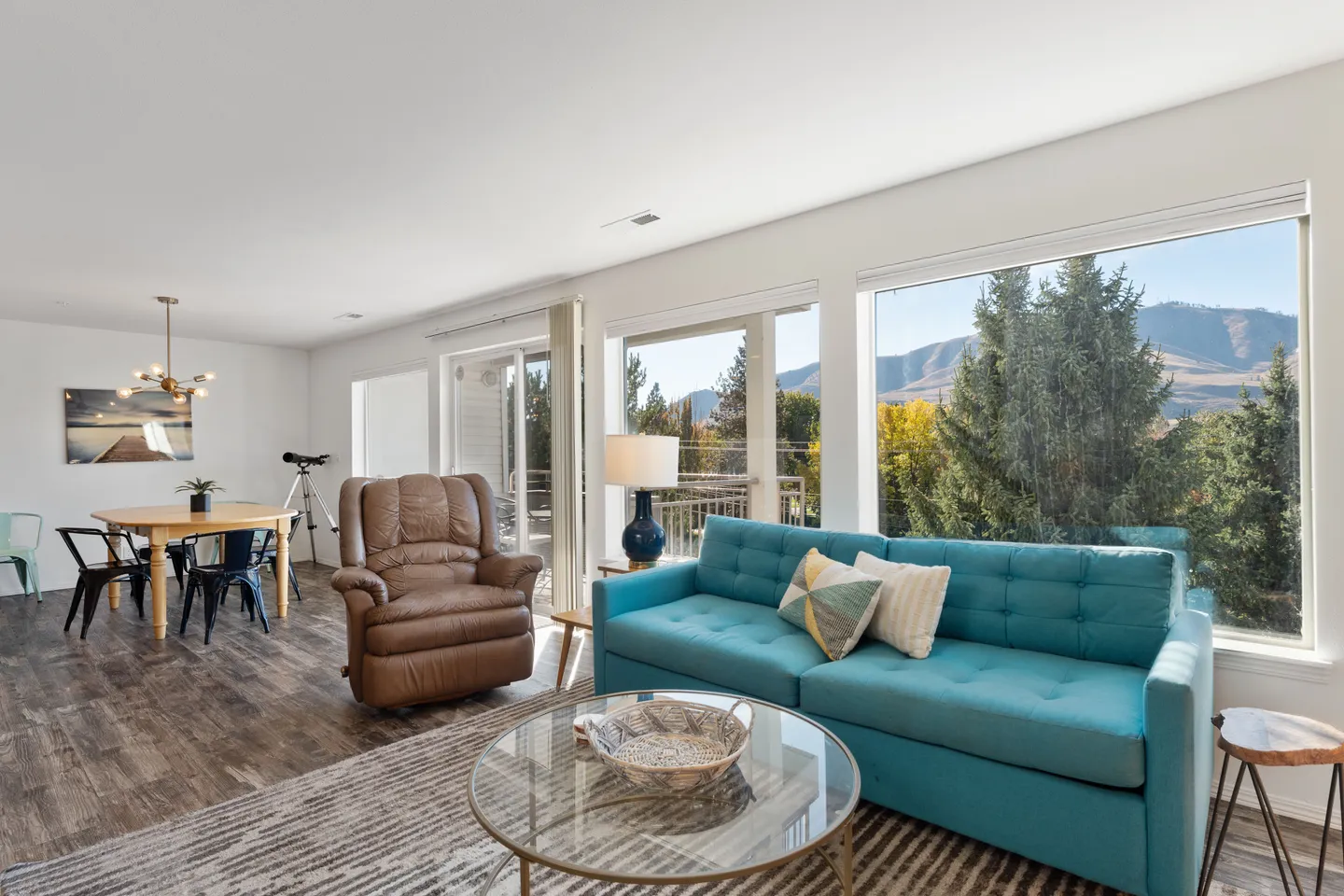 Living room with a teal sofa, brown recliner, round glass coffee table, and large windows overlooking trees and mountains.