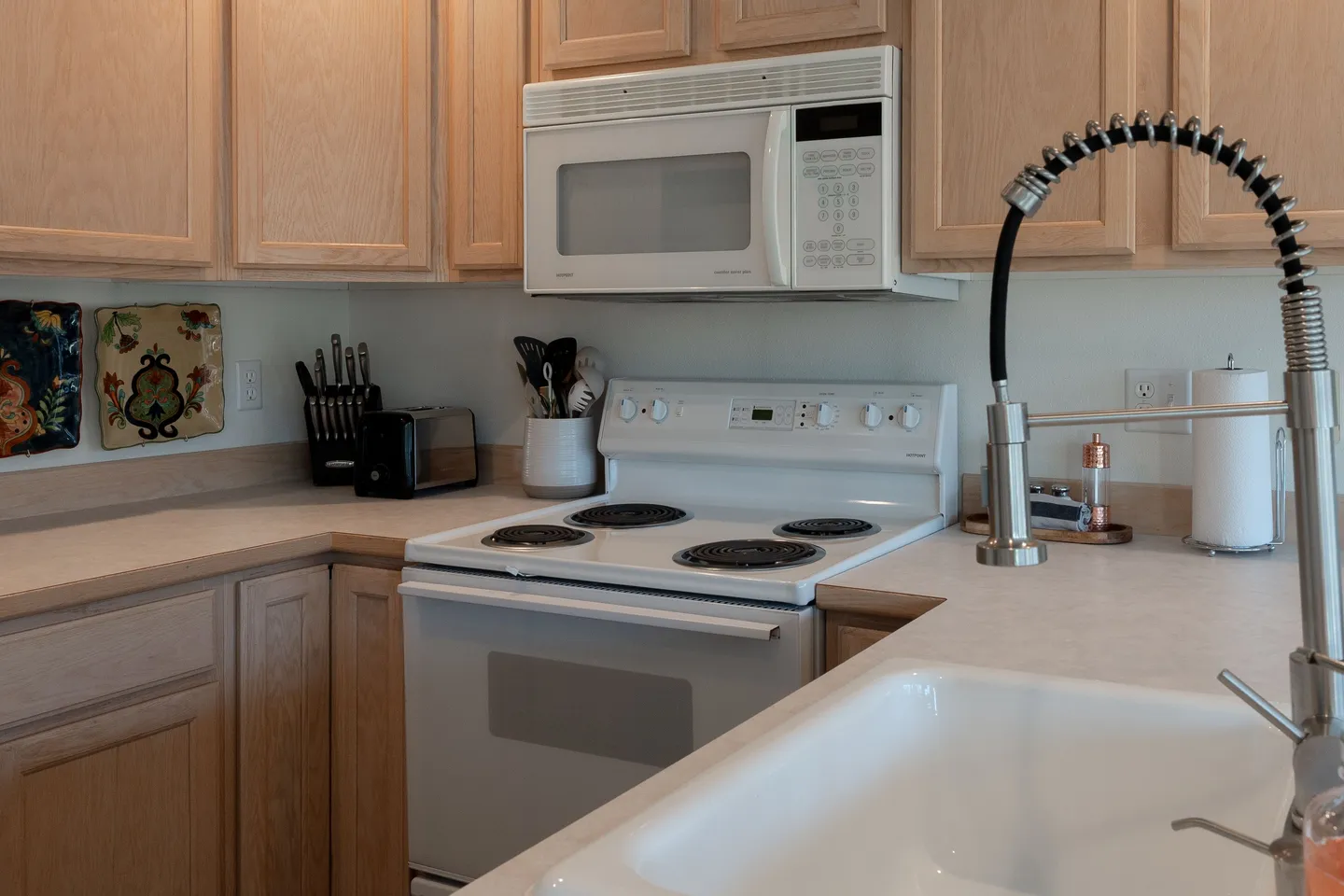 Kitchen counter area with an electric range, white over-the-range microwave, light wood cabinetry, and a high-arc faucet over a white sink.