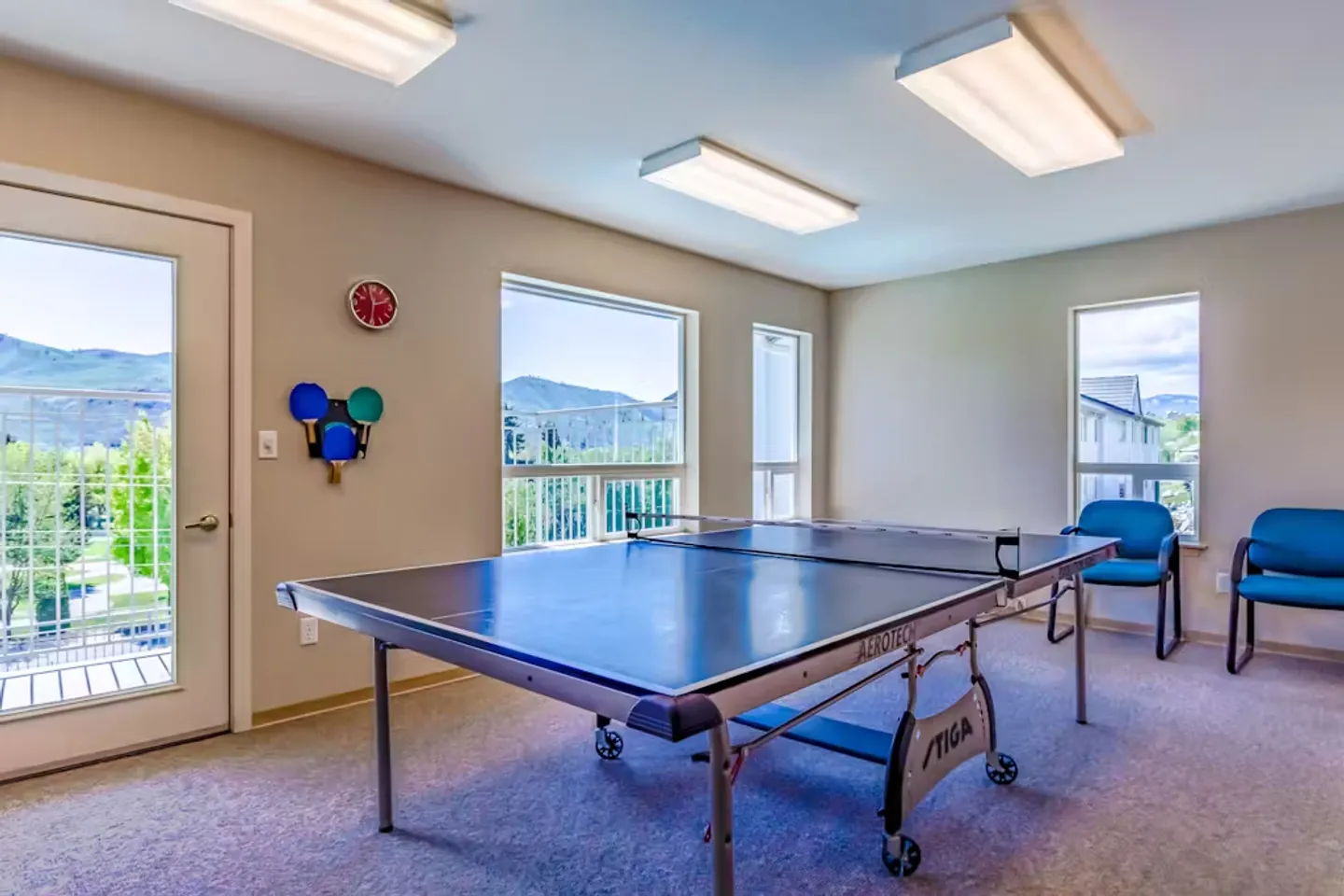 A brightly lit game room with carpeted flooring, featuring a ping-pong table and large windows overlooking mountains.