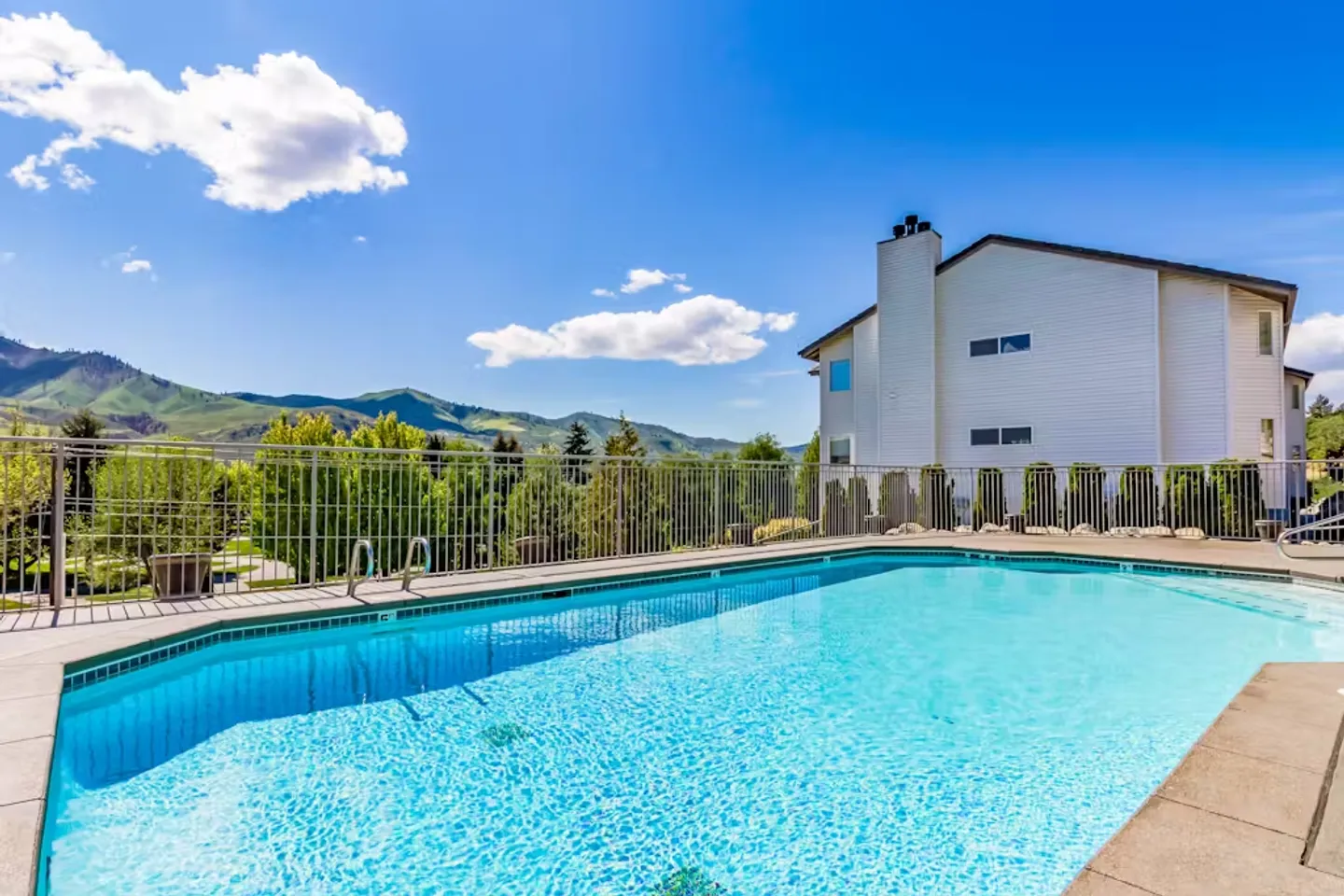 Outdoor swimming pool surrounded by a metal fence with views of green hills and a multi-story building under a sunny sky.