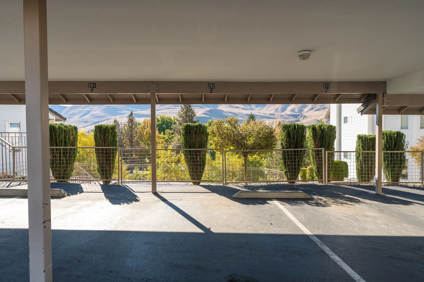 A covered parking carport looking out through a wire railing to manicured bushes, trees, and rolling hills in the background.