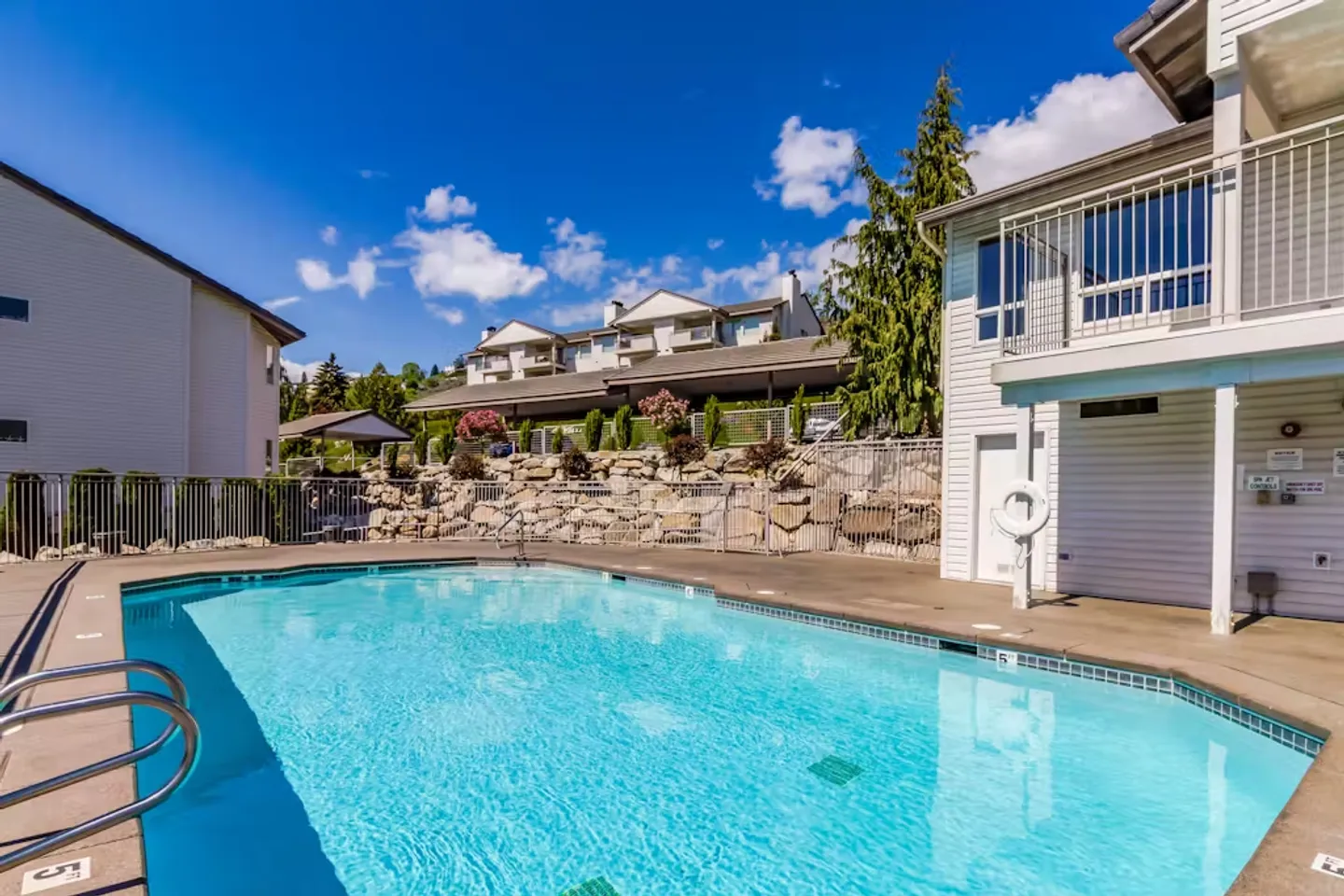 Outdoor swimming pool surrounded by a concrete patio, fence, and residential buildings under a blue sky.