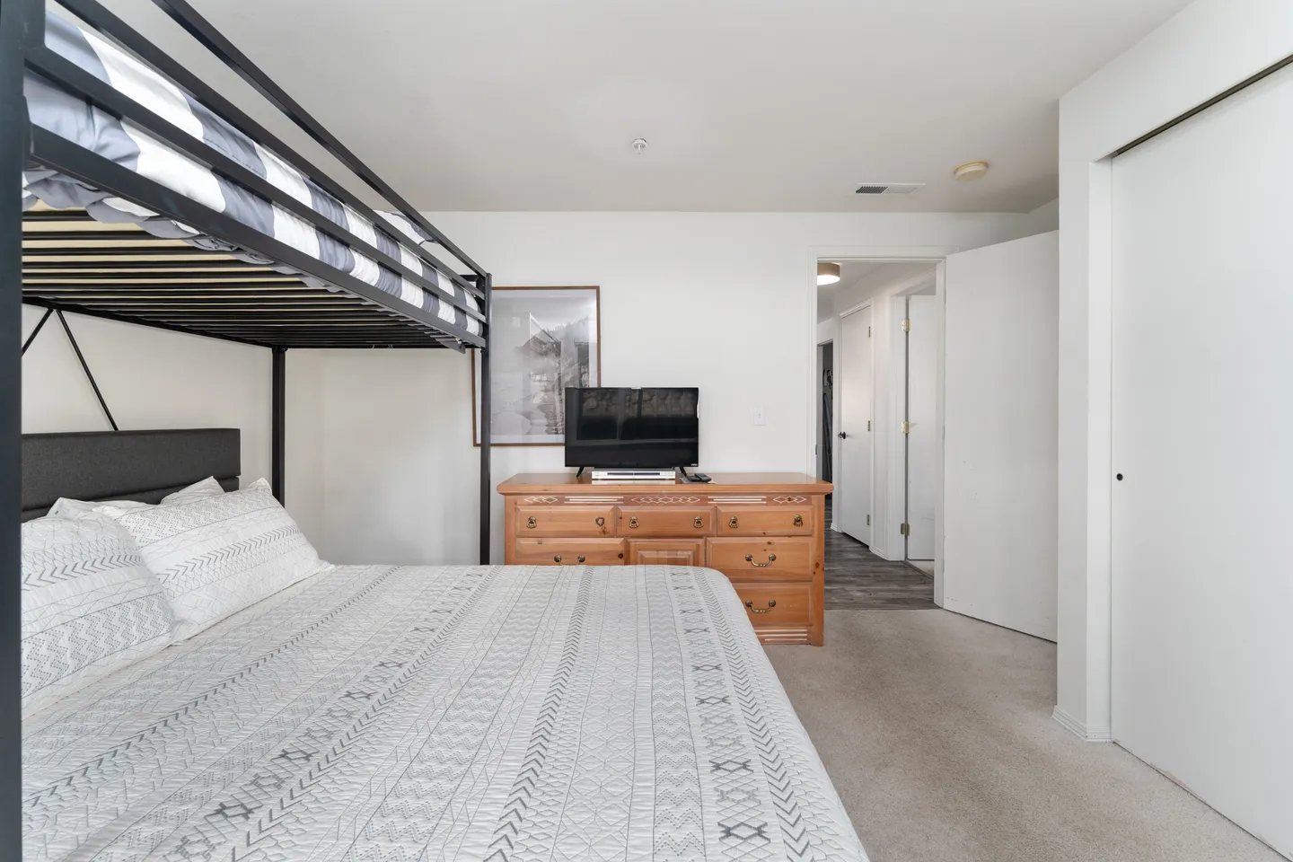 Bedroom with carpeted flooring, featuring a dark metal bunk bed, a wooden dresser with a TV on top, and a doorway leading to a hallway.